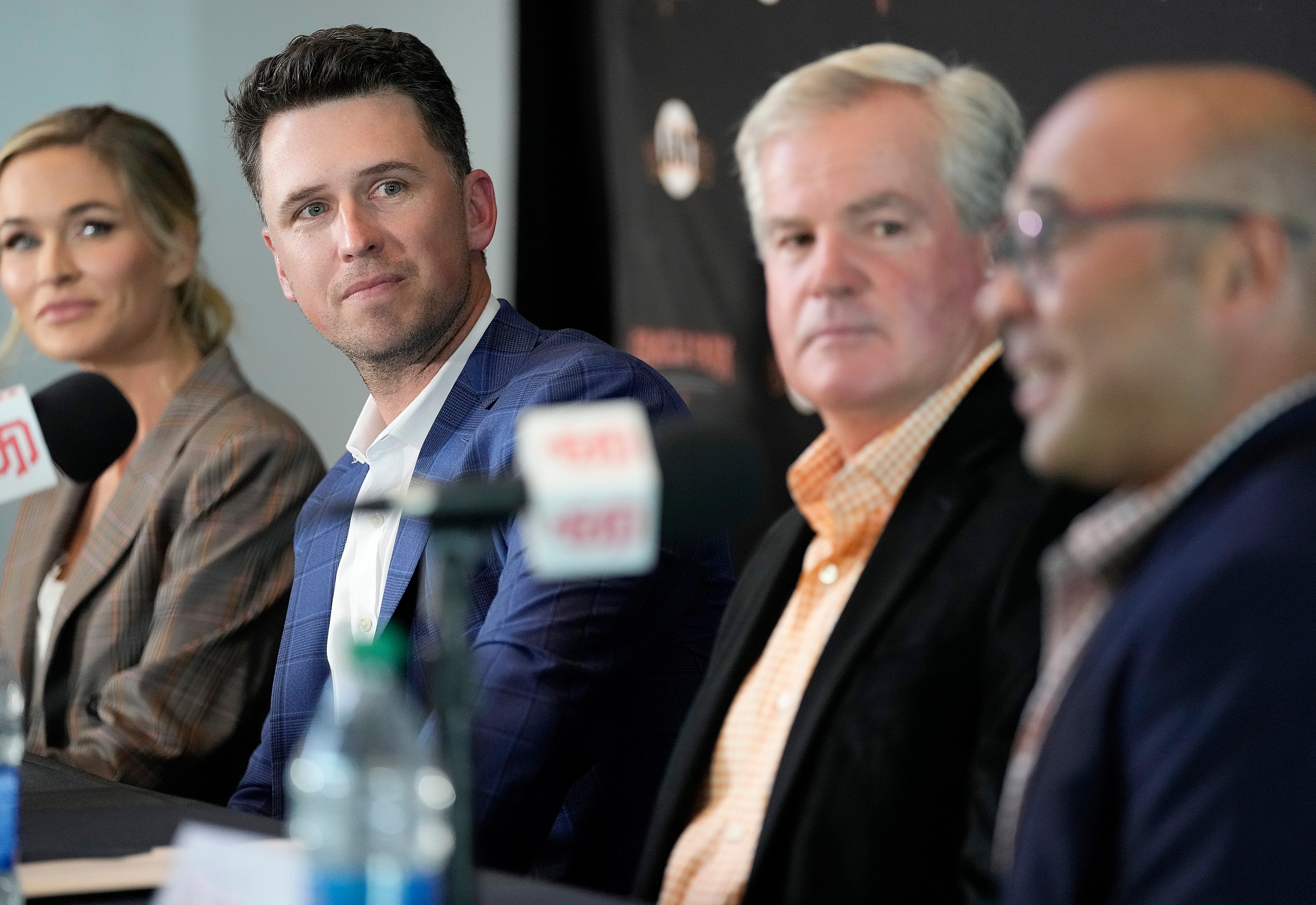 Four people in business attire sit at a table with microphones, engaged in a discussion. They wear serious expressions, and a water bottle is on the table.