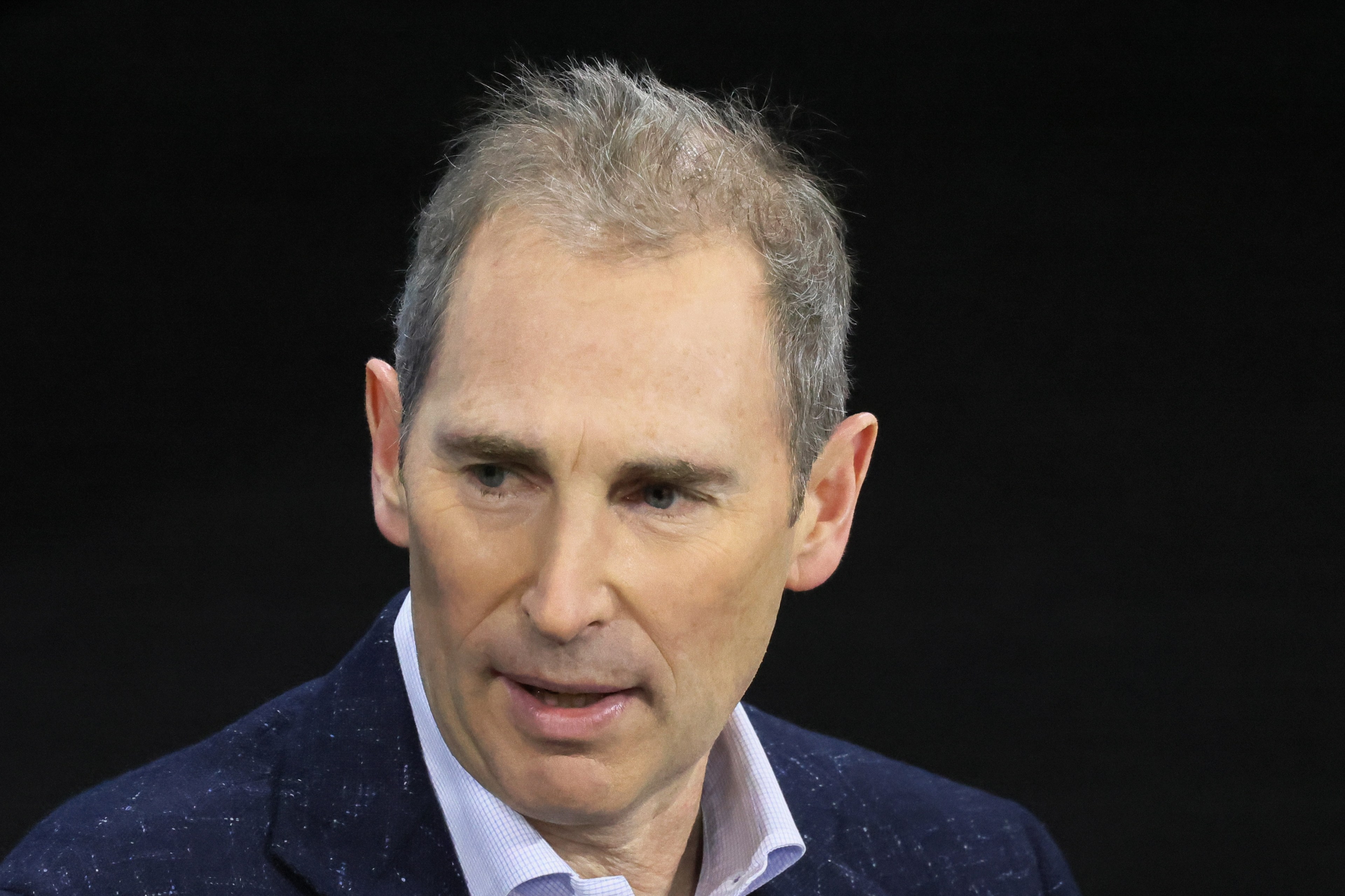 The image shows a man with short, graying hair and a suit jacket, looking slightly to the side against a dark background.