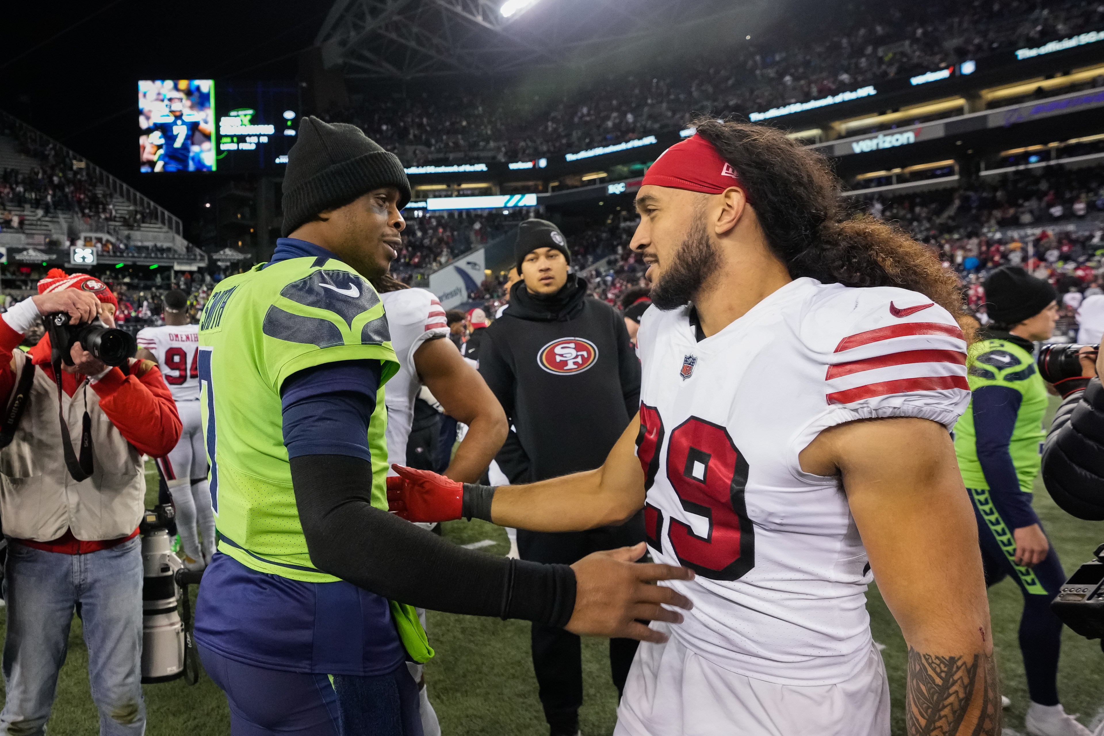 A football player in a green jersey and black beanie shakes hands with a player in a white jersey and red headband. They stand on a field with photographers nearby.