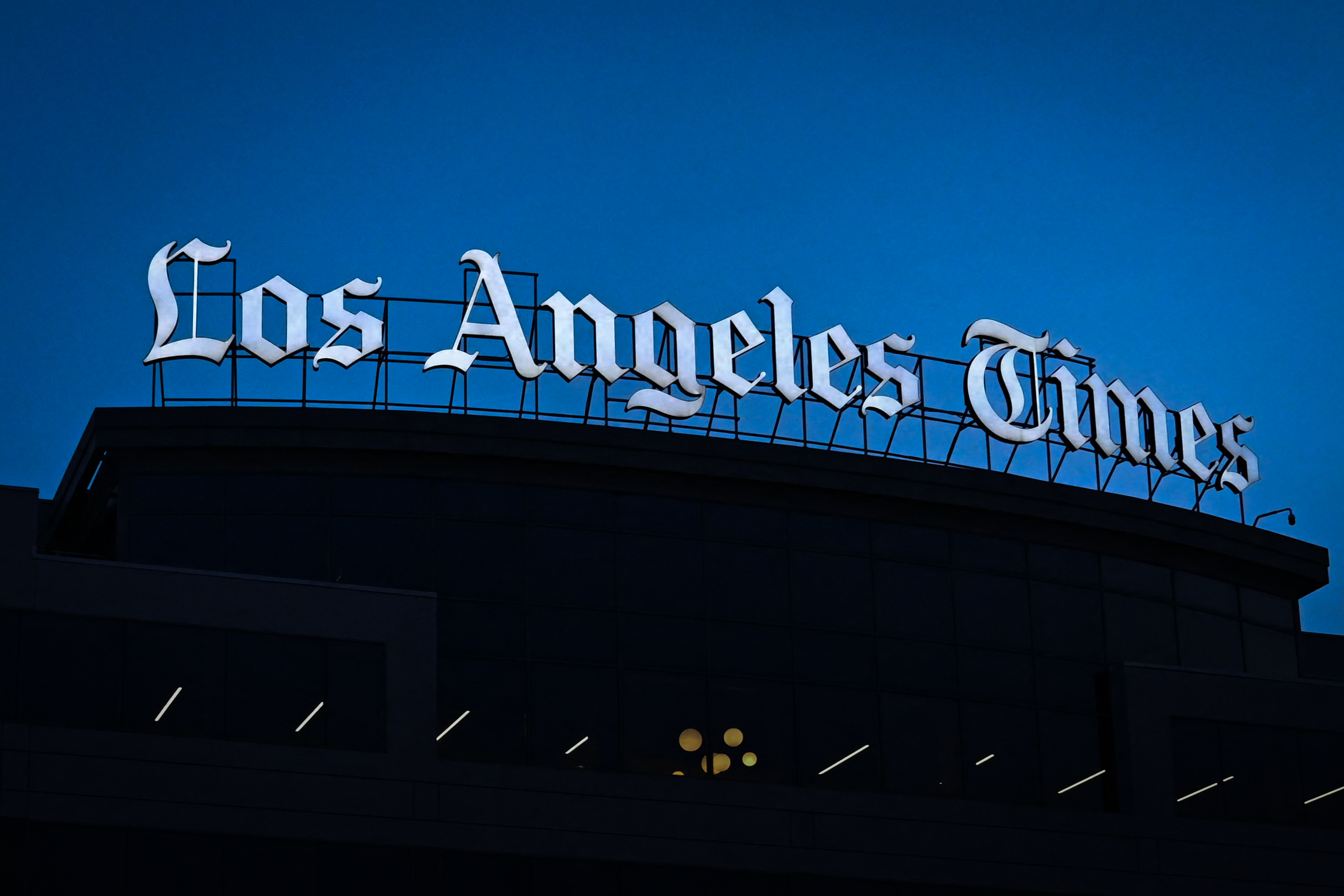 The image shows the "Los Angeles Times" sign, prominently displayed on a building. The sky behind is a deep blue, suggesting it is either dusk or dawn.