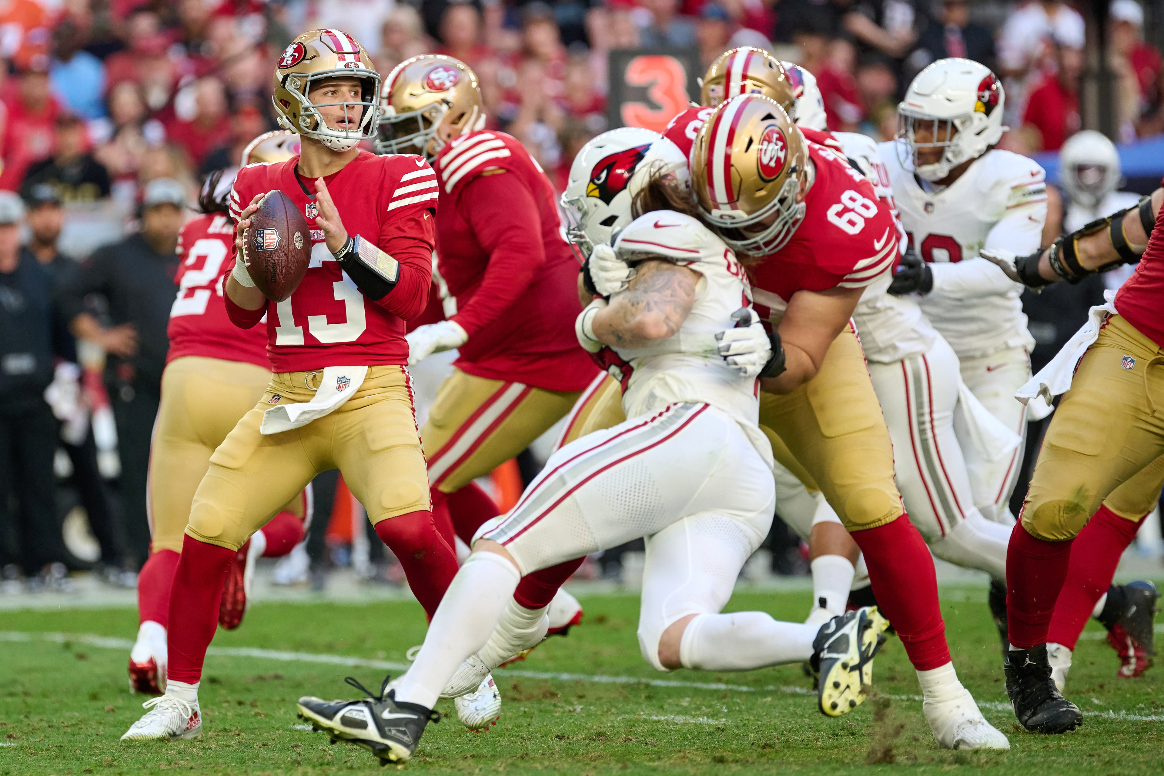 A football player in a red uniform, number 13, holds the ball, ready to throw, while teammates block players in white uniforms on a grassy field.