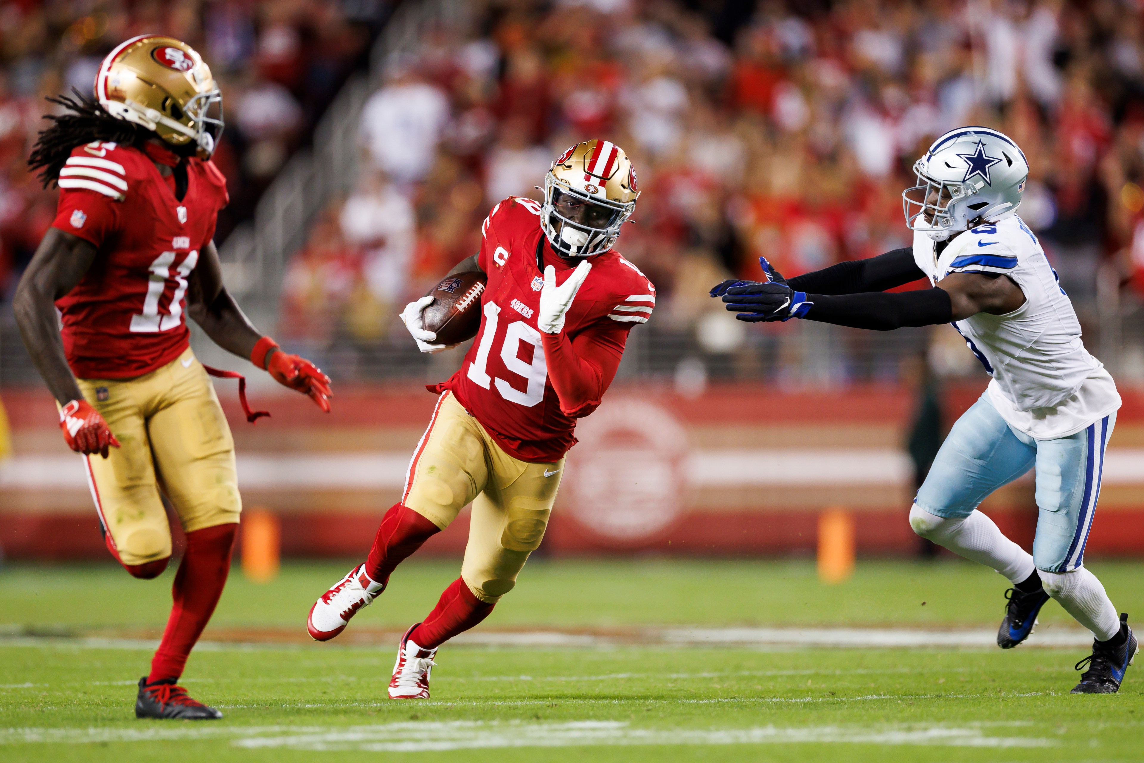 A football player in a red and gold uniform runs with the ball while an opponent in blue and white reaches out to tackle. Another teammate in red watches nearby.