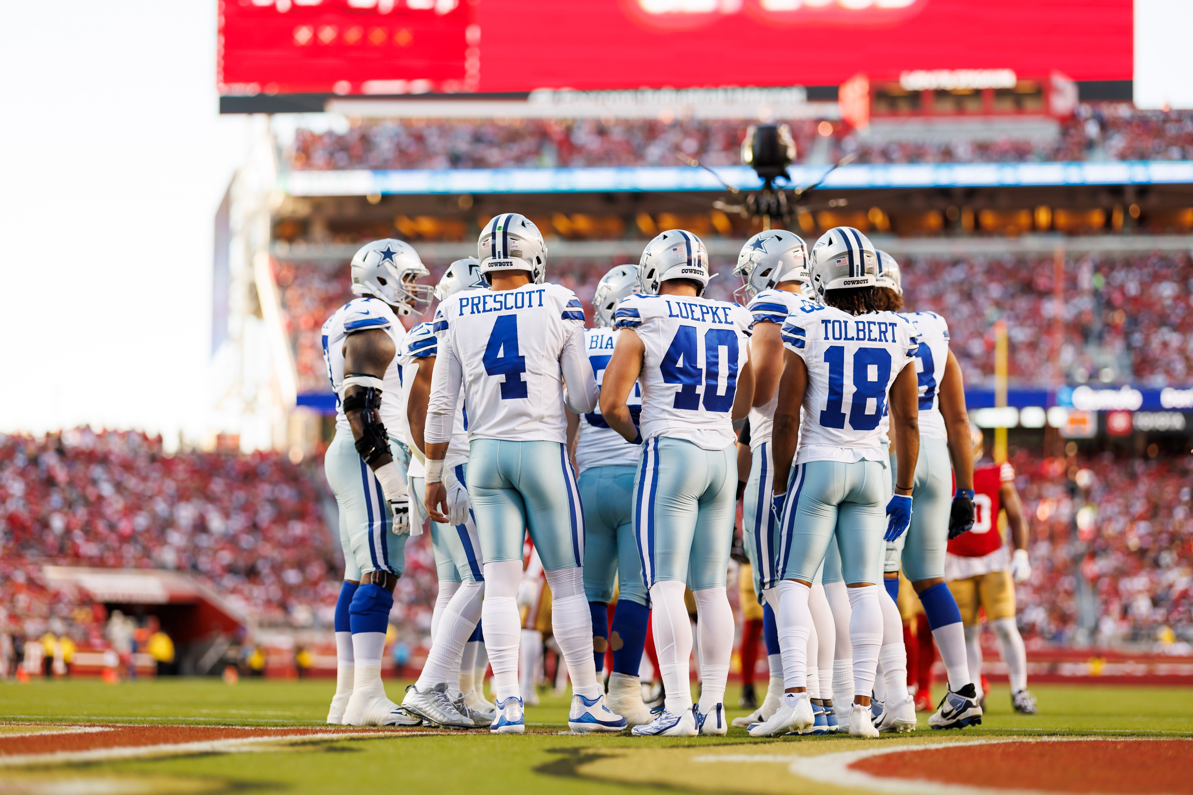 A group of American football players in blue and white uniforms huddle on a grassy field, with a large stadium and cheering crowd in the background.