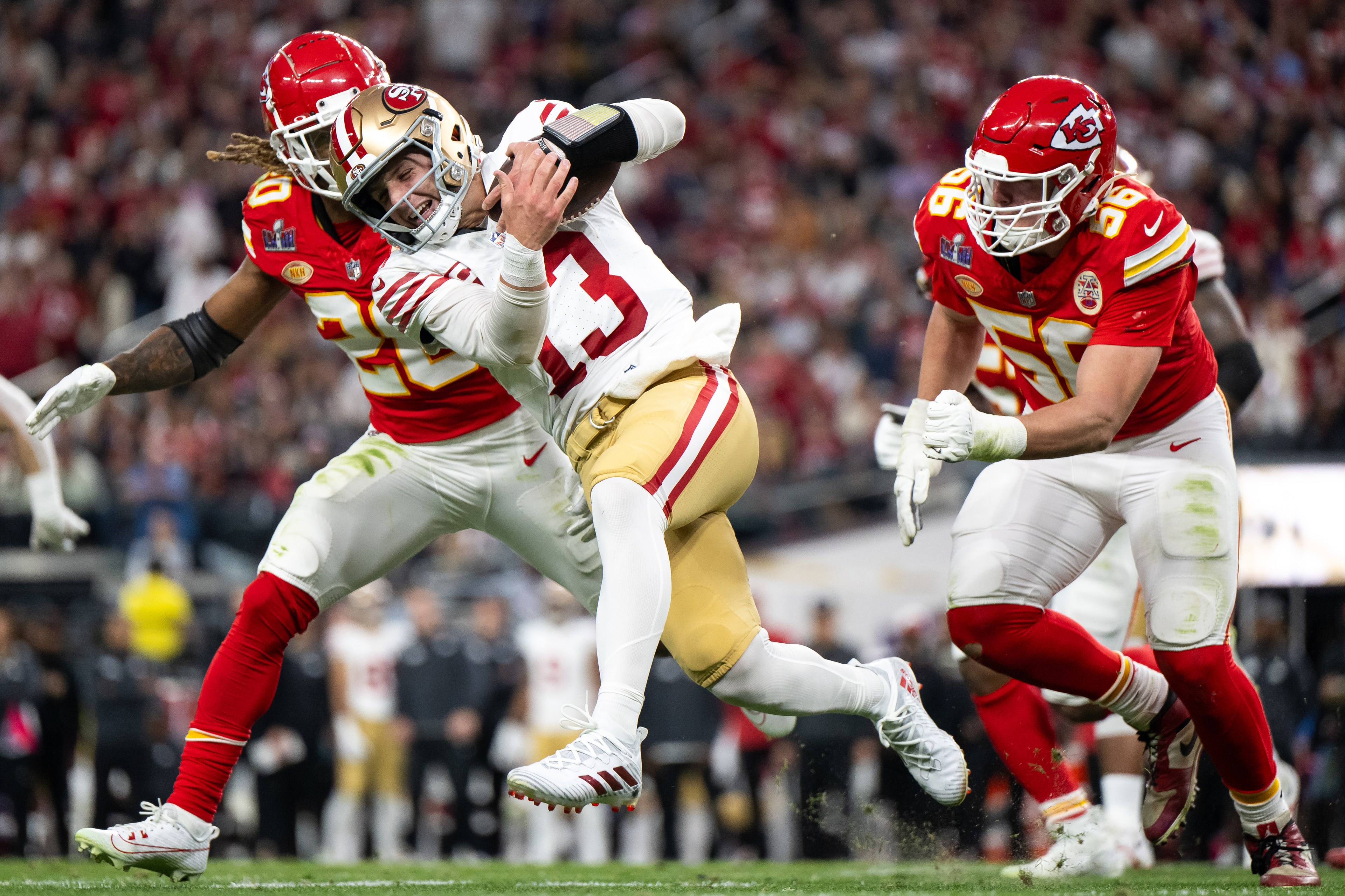 A football player in a white and gold uniform is running with the ball, being tackled by two opposing players in red uniforms during a game.