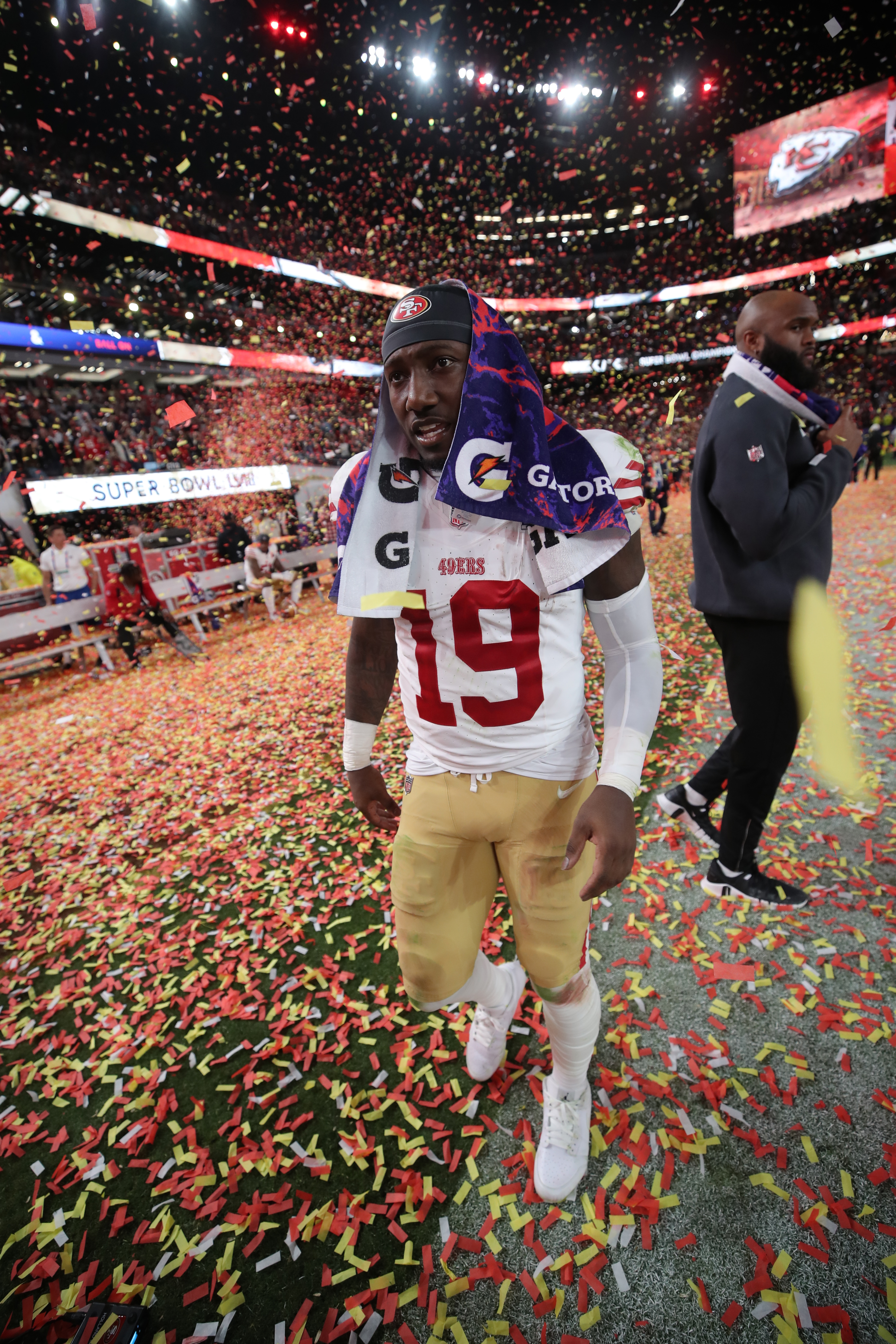 A football player wearing a 49ers uniform stands on a confetti-covered field with a Gatorade towel on his head, while celebrating people surround him.