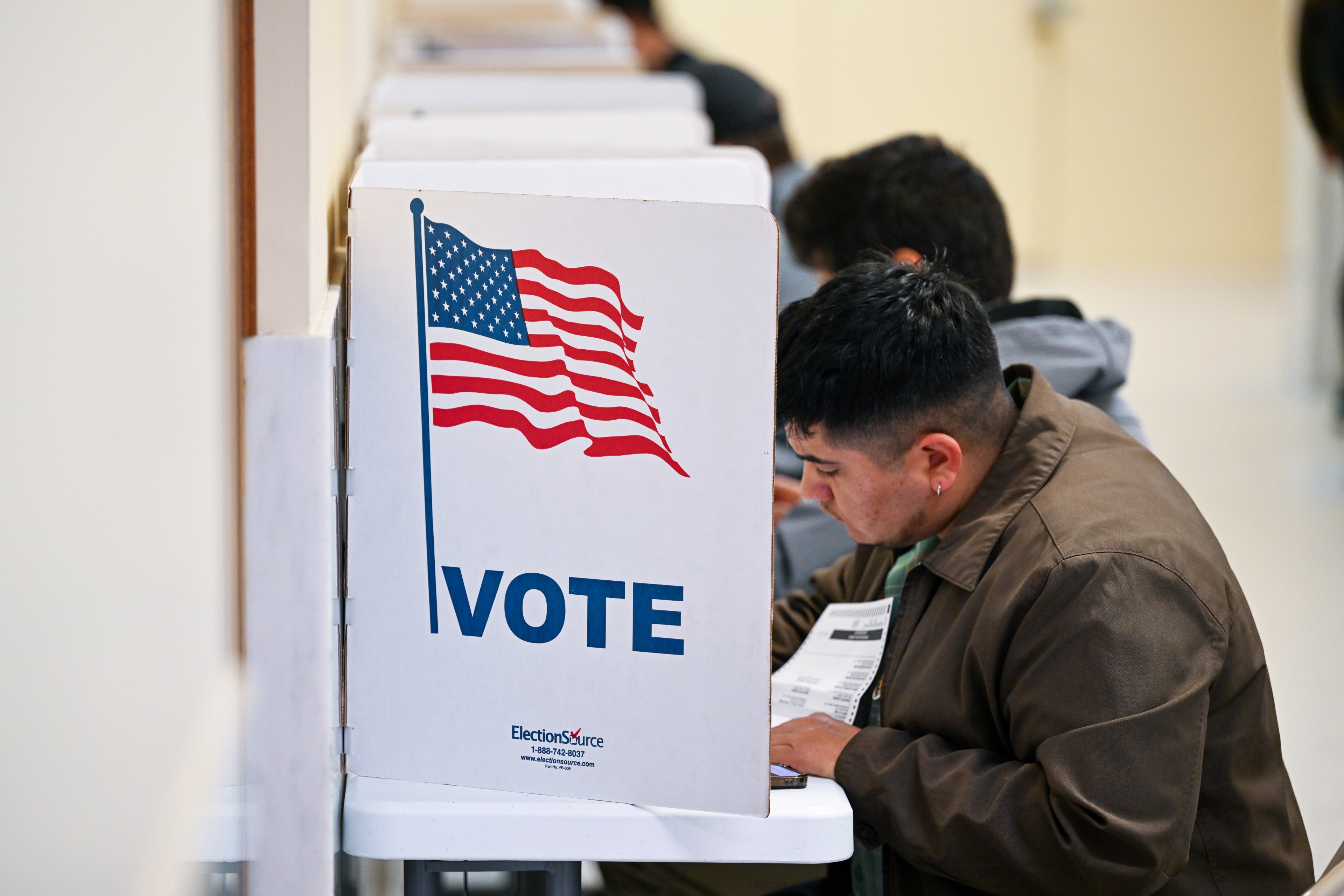 A man sits at a voting booth, filling out a ballot. The booth features an American flag and the word "VOTE" in bold letters. Other voters are visible in the background.