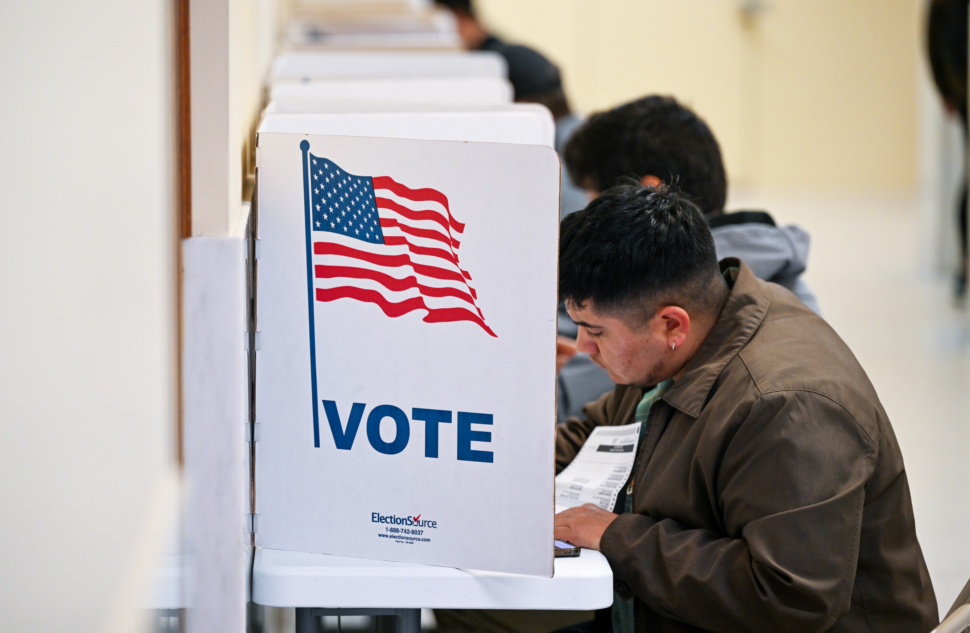 A man sits at a voting booth, filling out a ballot. The booth features an American flag and the word "VOTE" in bold letters. Other voters are visible in the background.