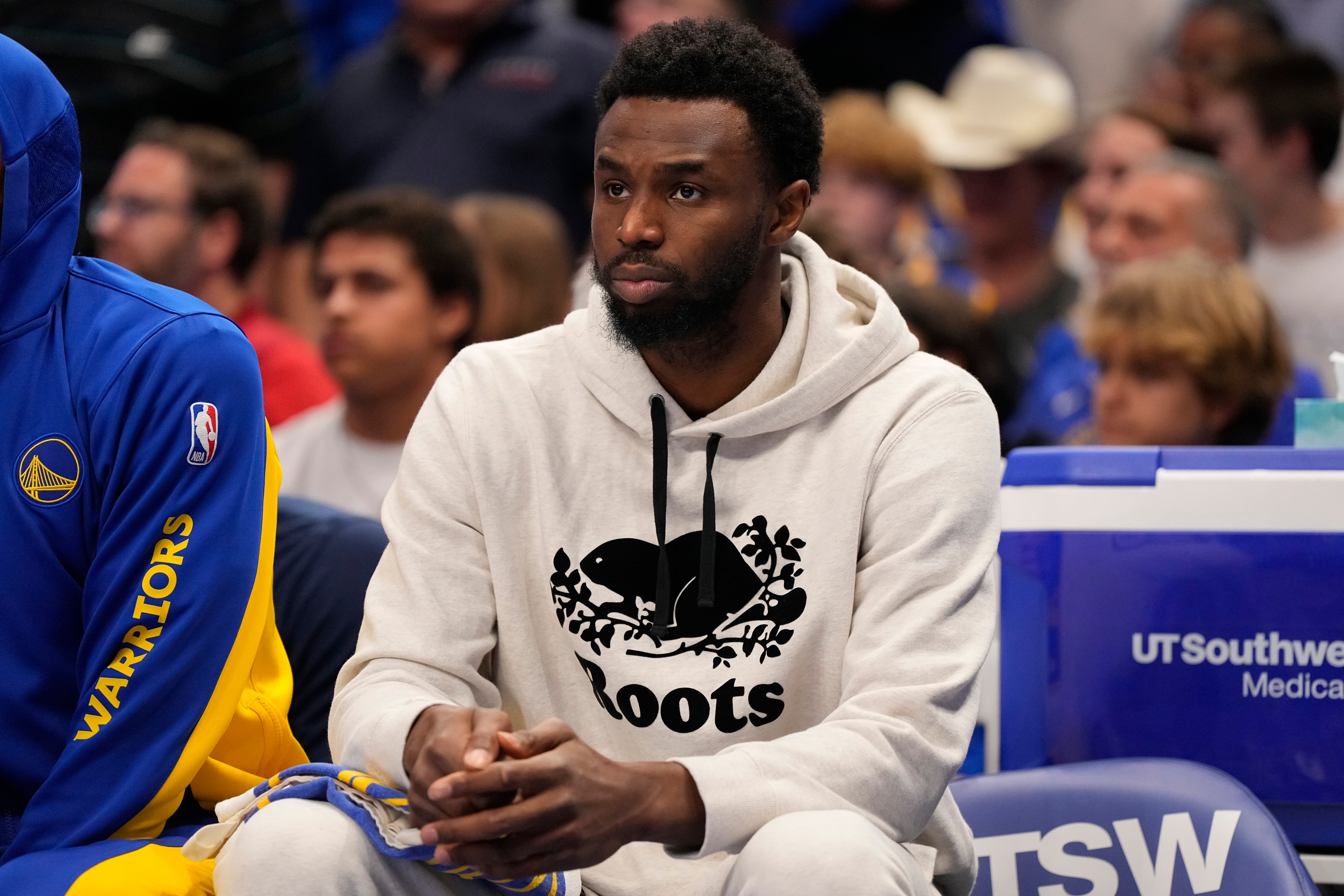 A man sits on a sports bench wearing a white &quot;Roots&quot; hoodie, looking thoughtful. The surrounding crowd and a person in a blue Warriors hoodie are visible.