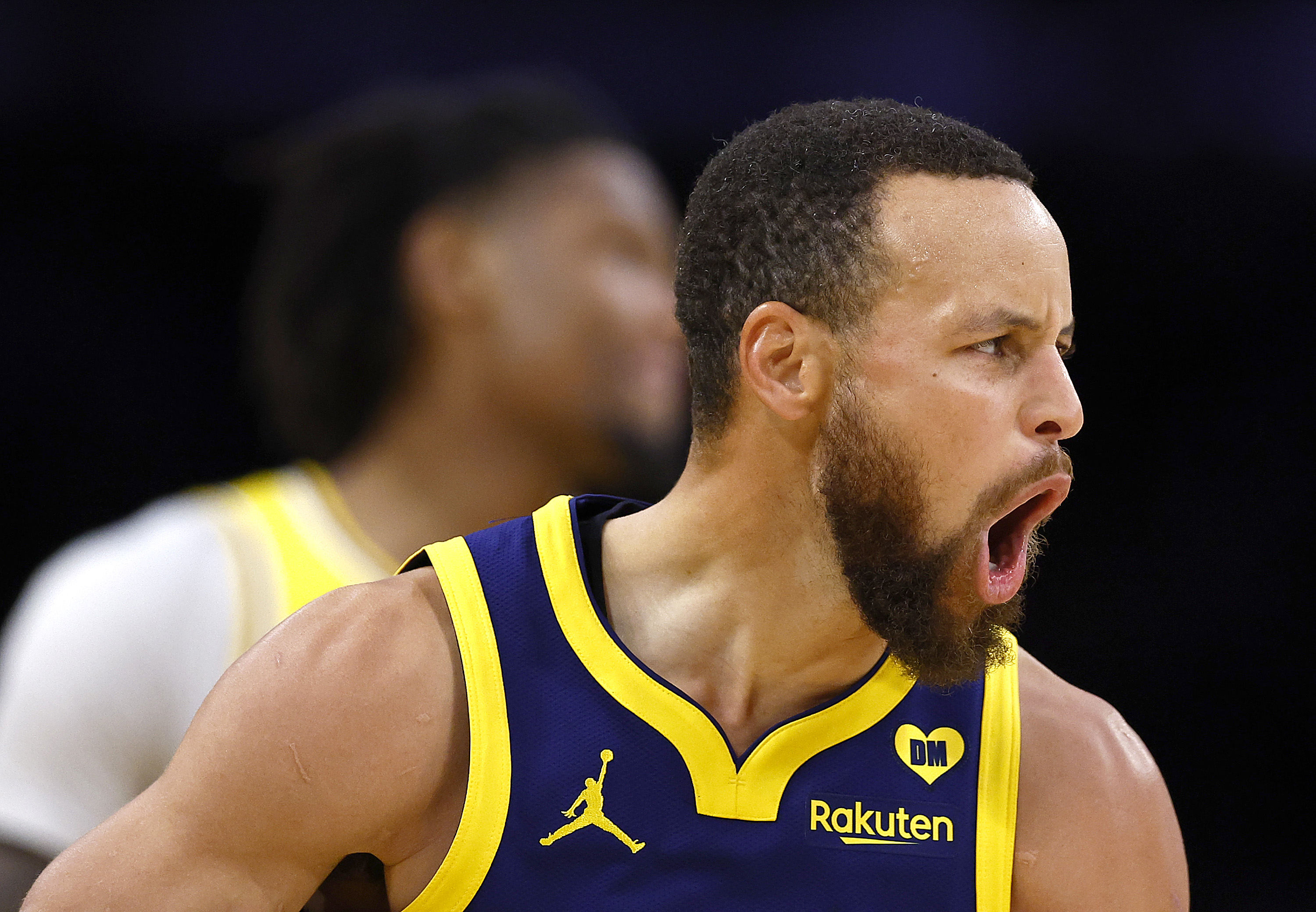 A basketball player in a blue and yellow jersey with "Rakuten" is enthusiastically yelling. Another player is blurred in the background.