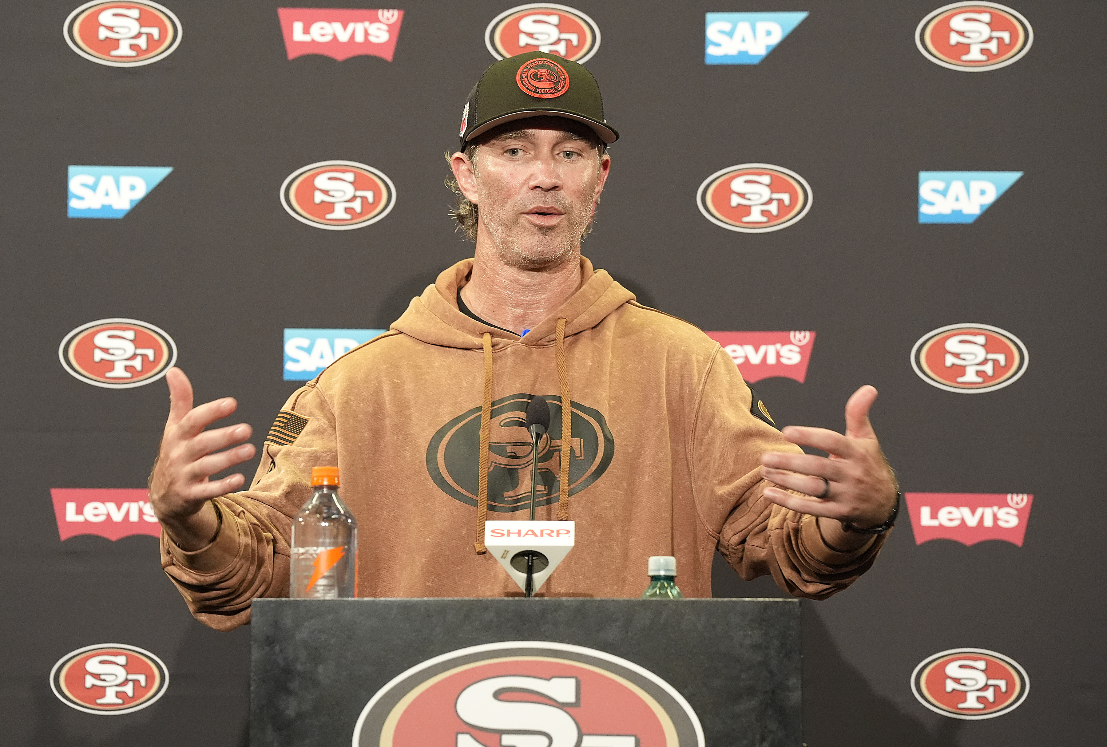 A person in a hoodie and cap speaks at a podium with sports logos and brand logos in the background, including SAP and Levi's. There are beverage bottles on the podium.