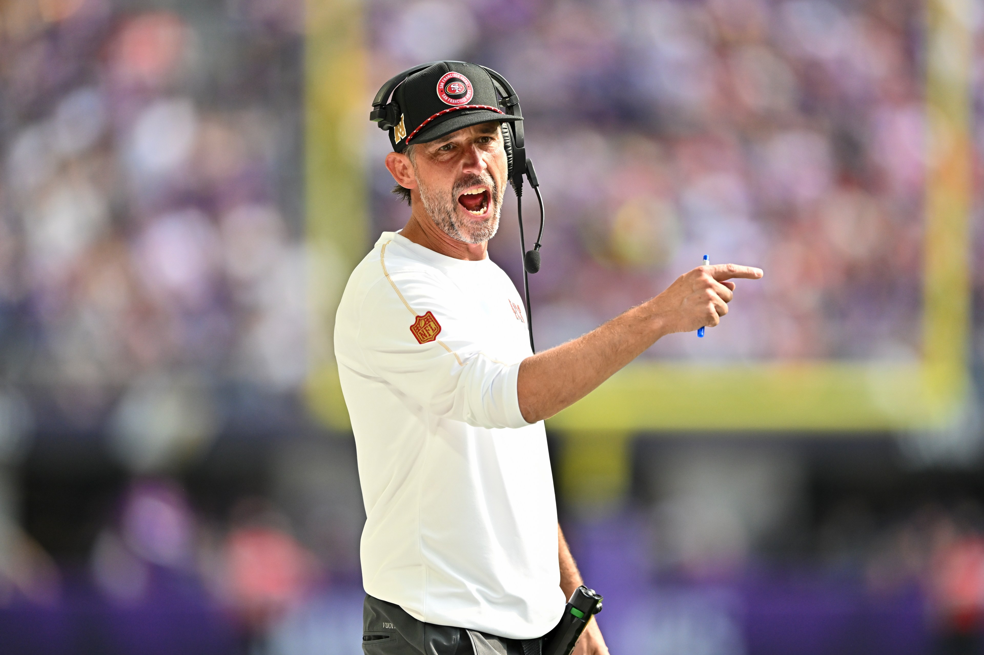 A man wearing a headset and cap is passionately gesturing and pointing with a pen. He is dressed in a white shirt, possibly during a sports event.