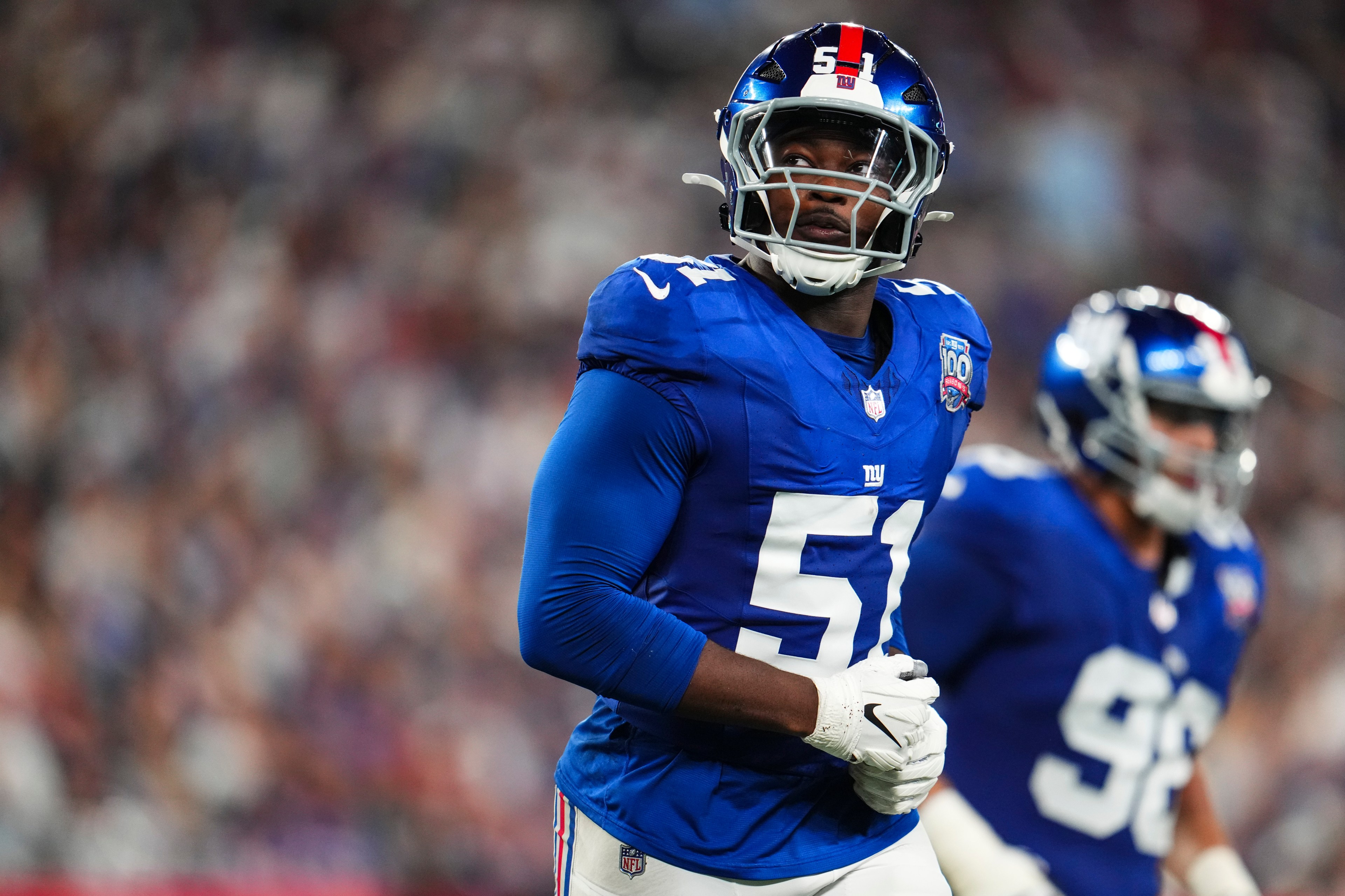 A football player in a blue jersey with the number 51 is on the field, wearing a helmet and gloves. Another player is in the background, slightly out of focus.