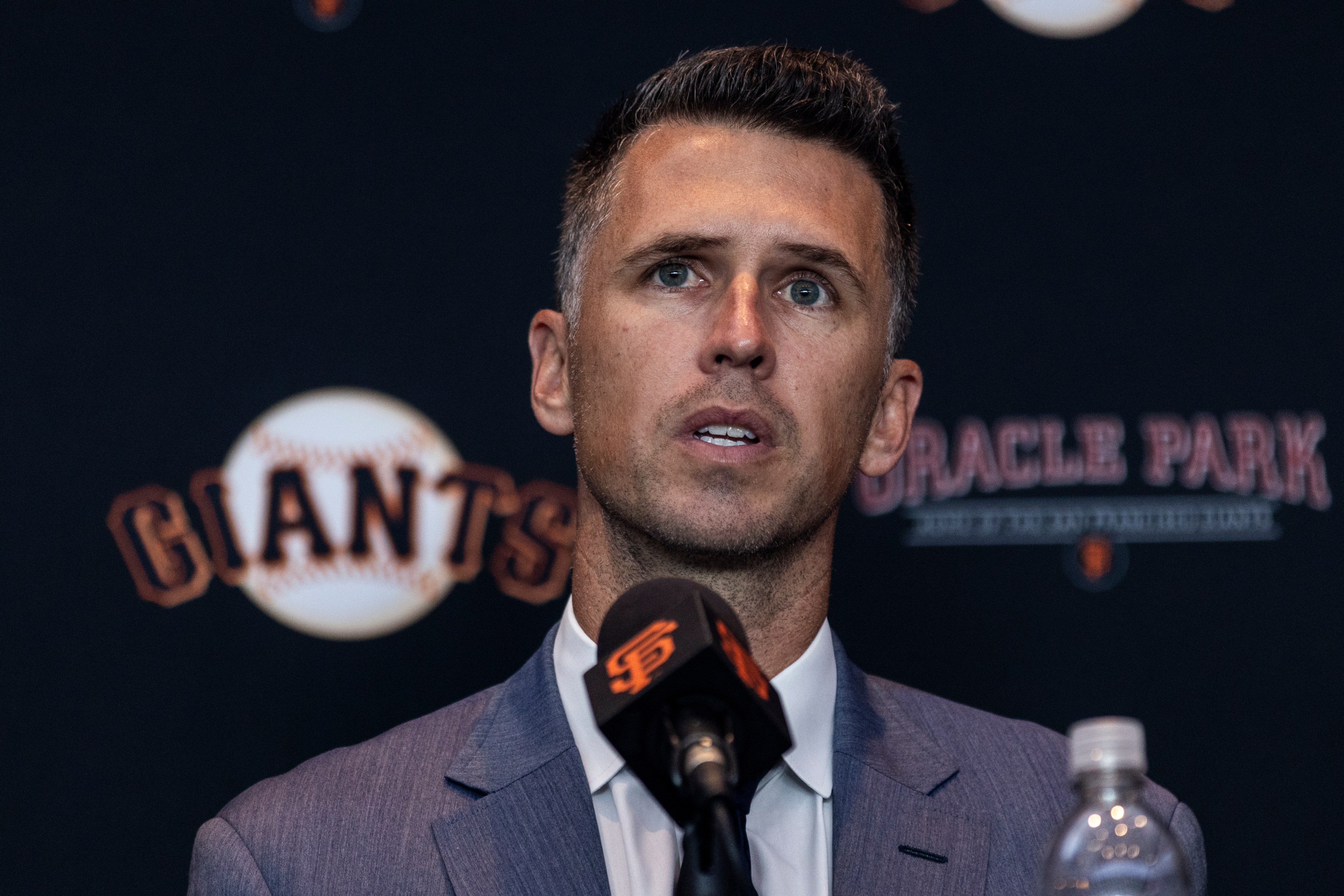 A man in a suit speaks at a microphone with the San Francisco Giants logo. The backdrop shows the words "Giants" and "Oracle Park" prominently.