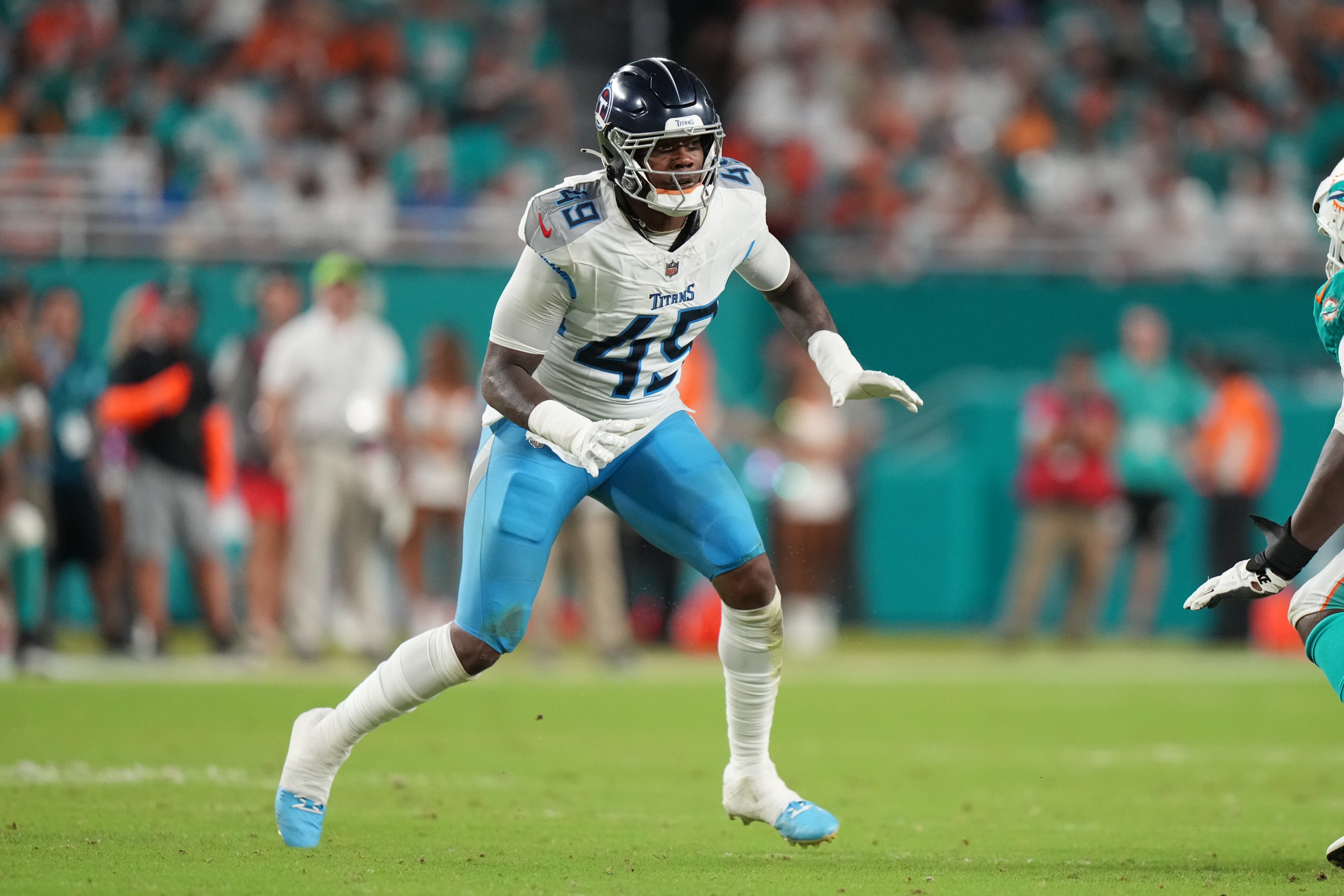 A football player in a white and blue uniform is in action on the field, with an opposing player partially visible. The background shows a blurred crowd.