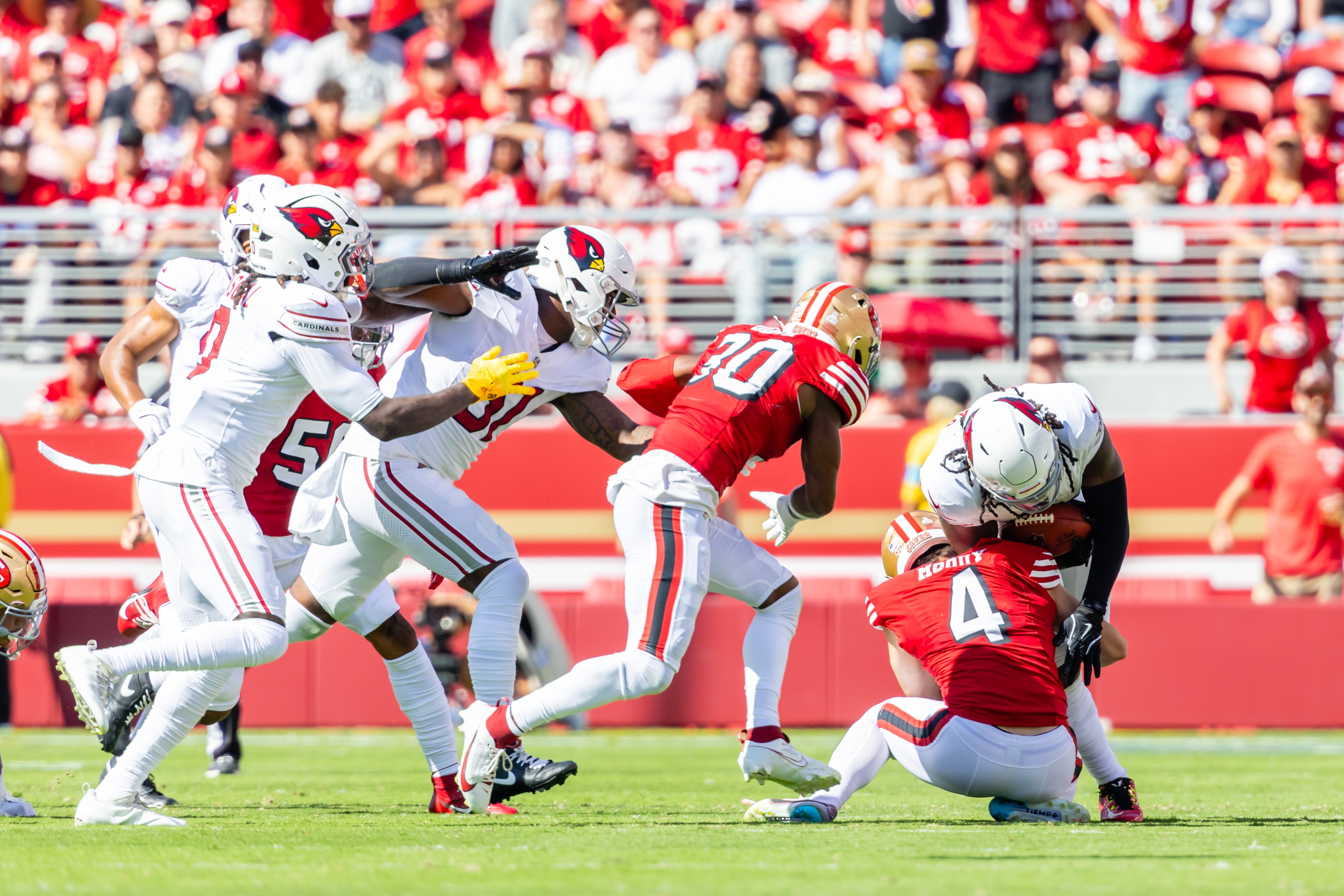 In the image, football players in red and white jerseys are in action. One player in red is tackling a player with a football, while others move toward the play.