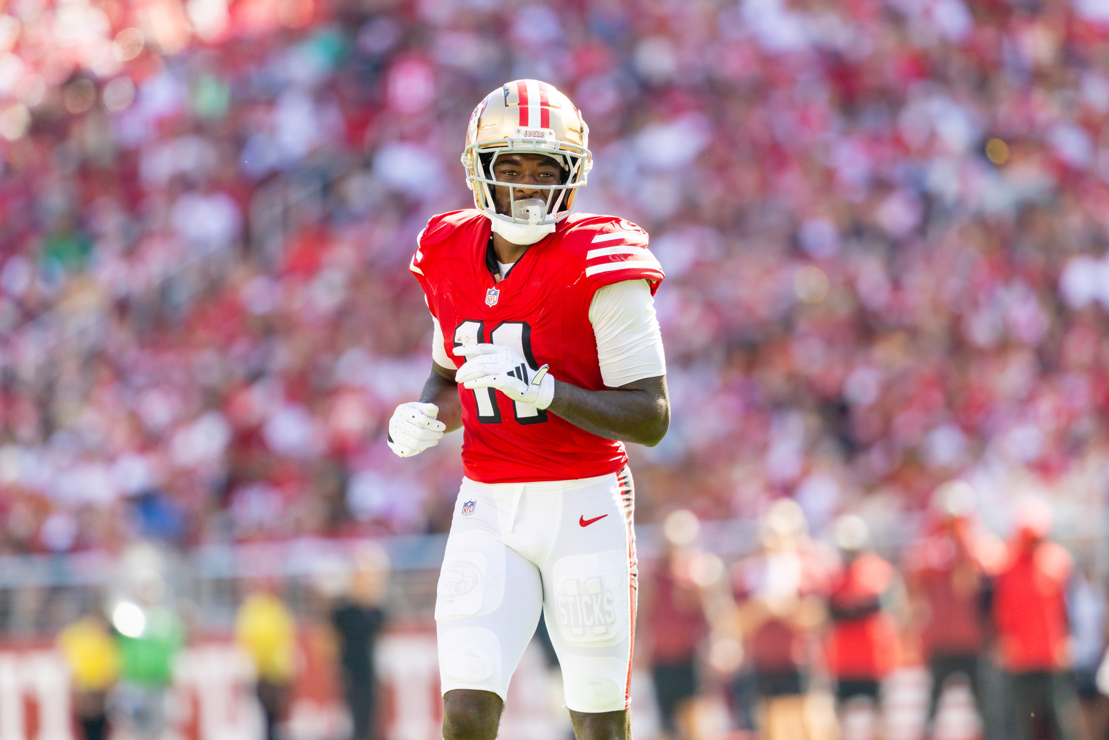 A football player in a red jersey with the number 11 runs on a field. He's wearing a helmet and white pants, with a crowd blurred in the background.