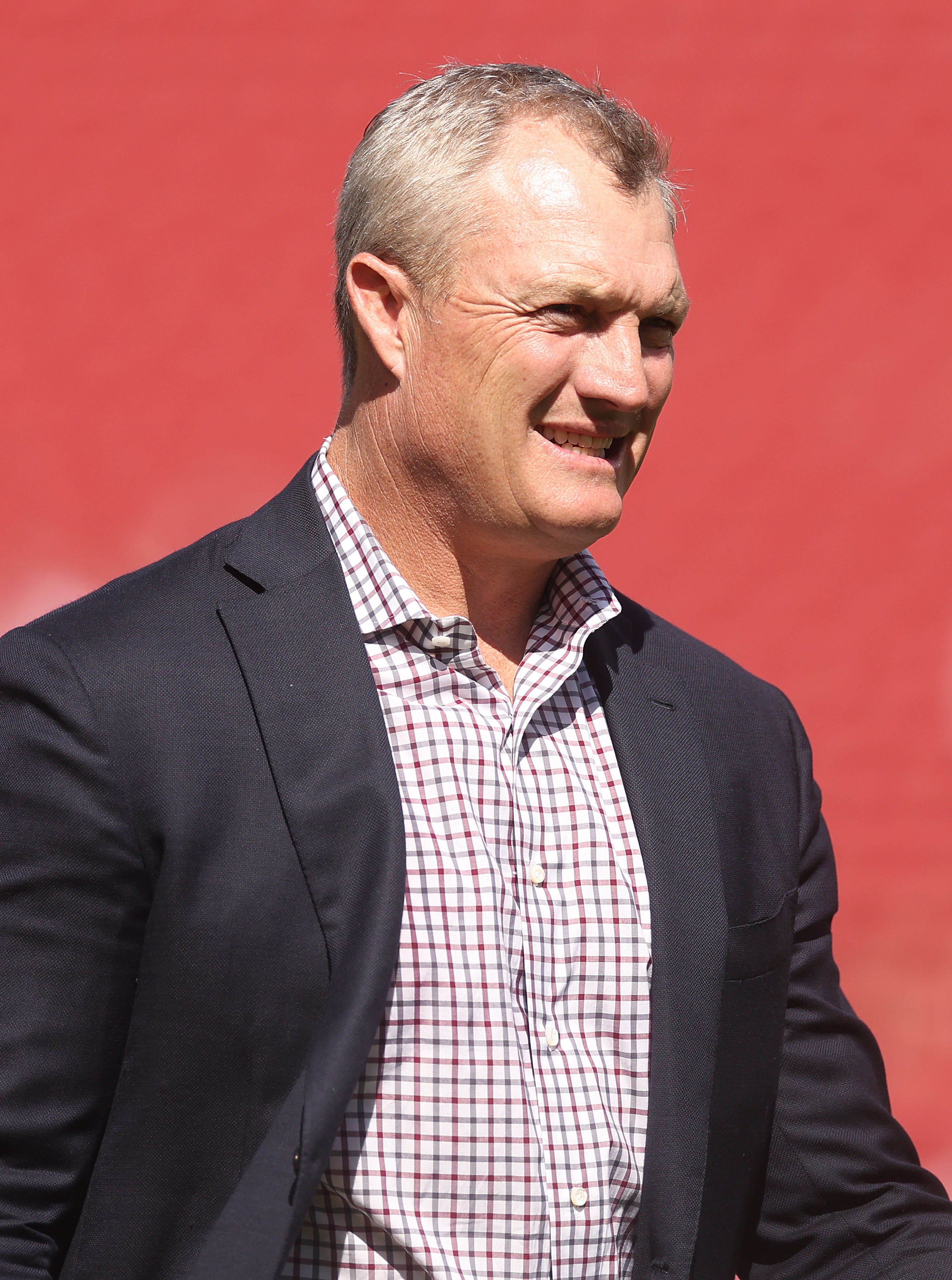 A smiling man with short gray hair is wearing a dark suit jacket over a checkered shirt, standing against a solid red background.