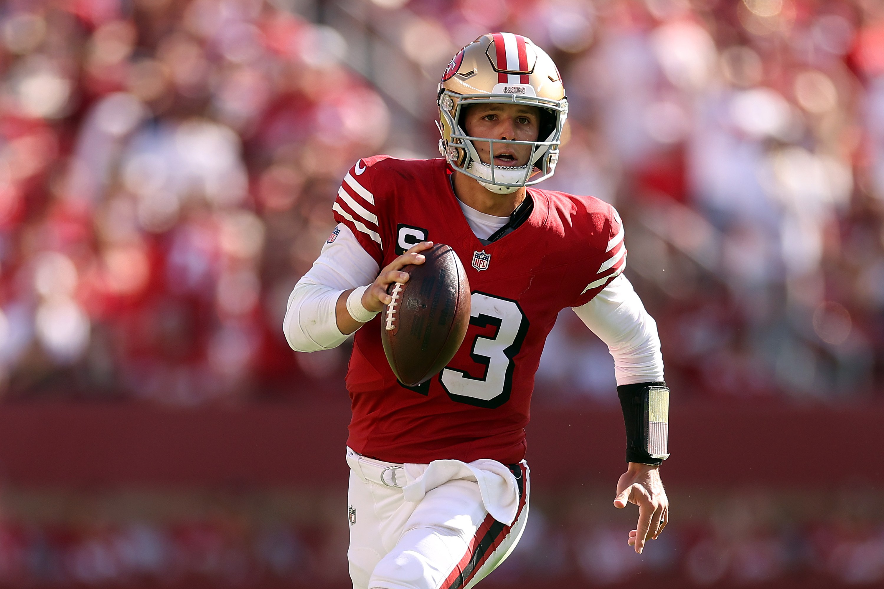 A football player in a red and gold uniform is running on a field, holding a football in his right hand. The background shows a crowd in a stadium.