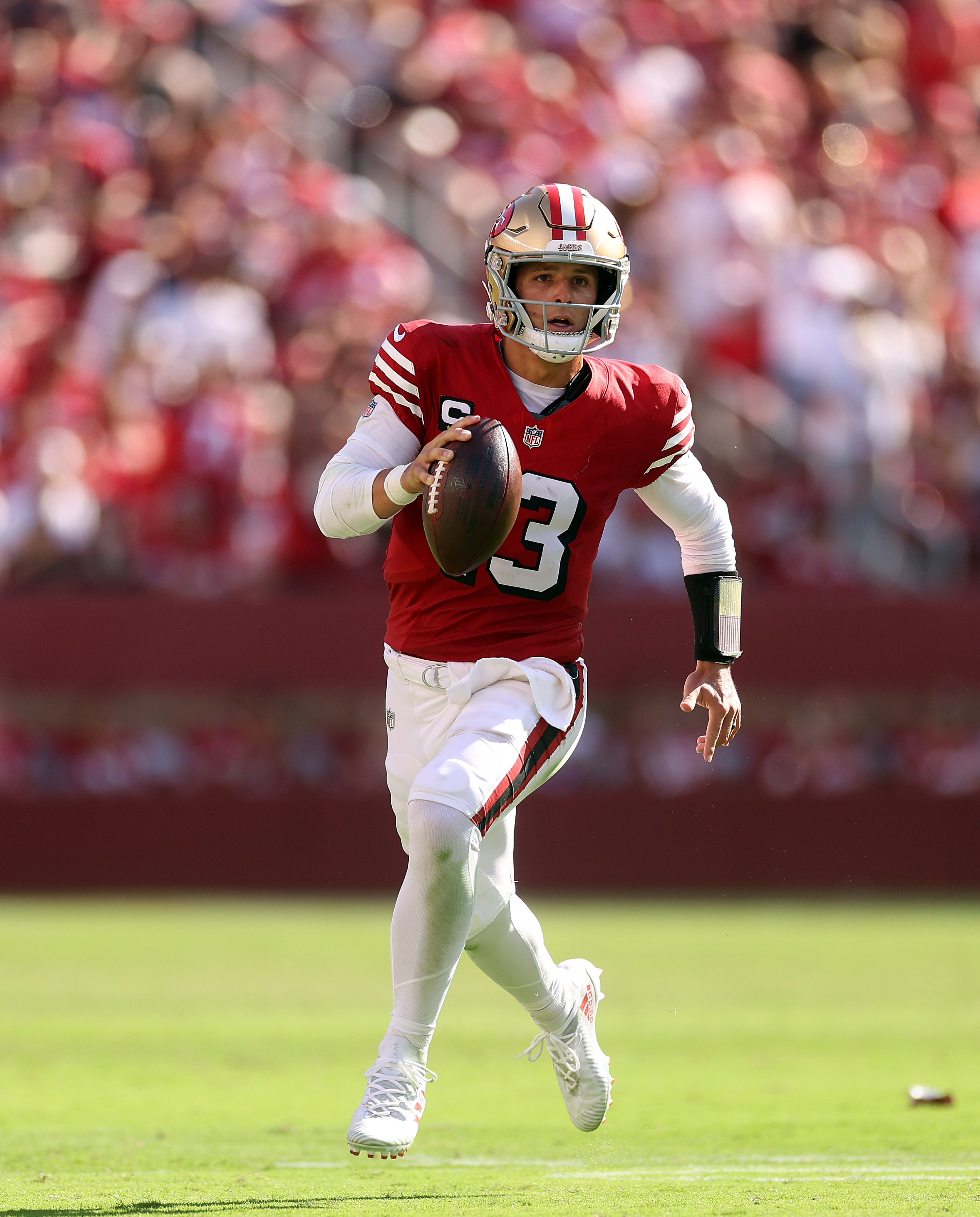 A football player in a red and gold uniform is running on a field, holding a football in his right hand. The background shows a crowd in a stadium.