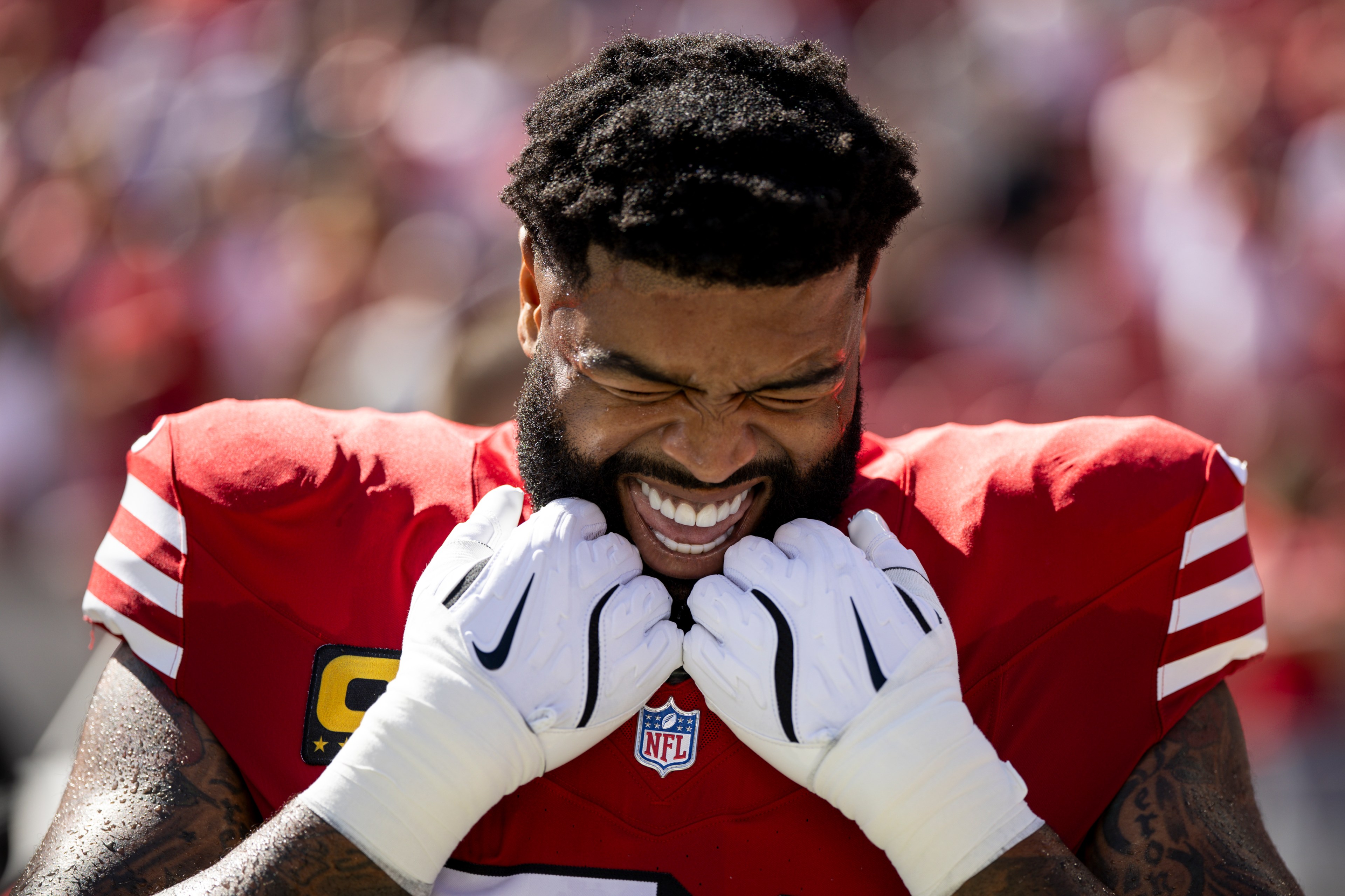 A football player in a red jersey and white gloves stands with eyes closed, teeth clenched, and fists near his face, seeming determined before a game.