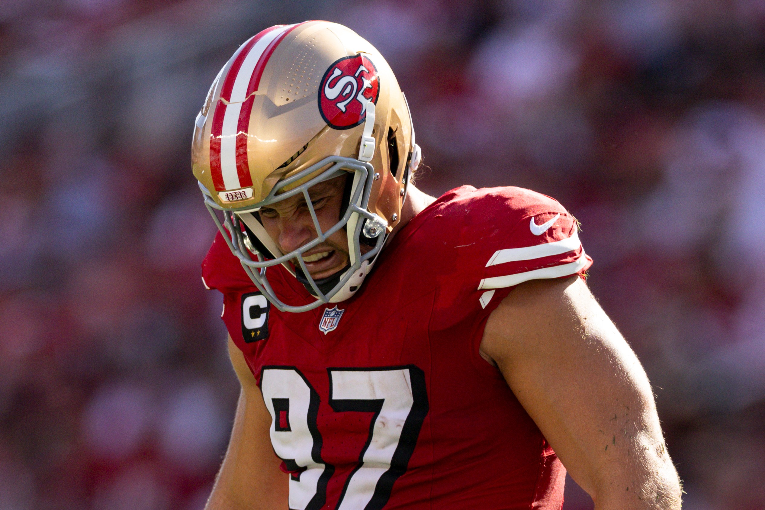 A football player wearing a red jersey and white pants, marked with number 97, stands on the field in action, sporting a gold helmet with a team logo.