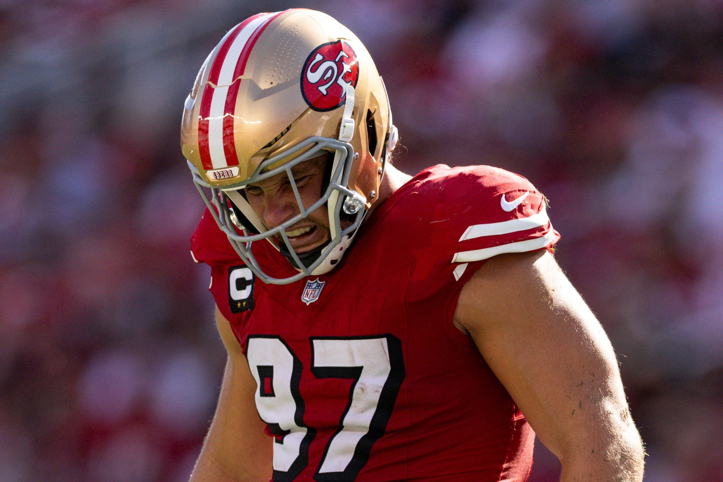 A football player wearing a red jersey and white pants, marked with number 97, stands on the field in action, sporting a gold helmet with a team logo.