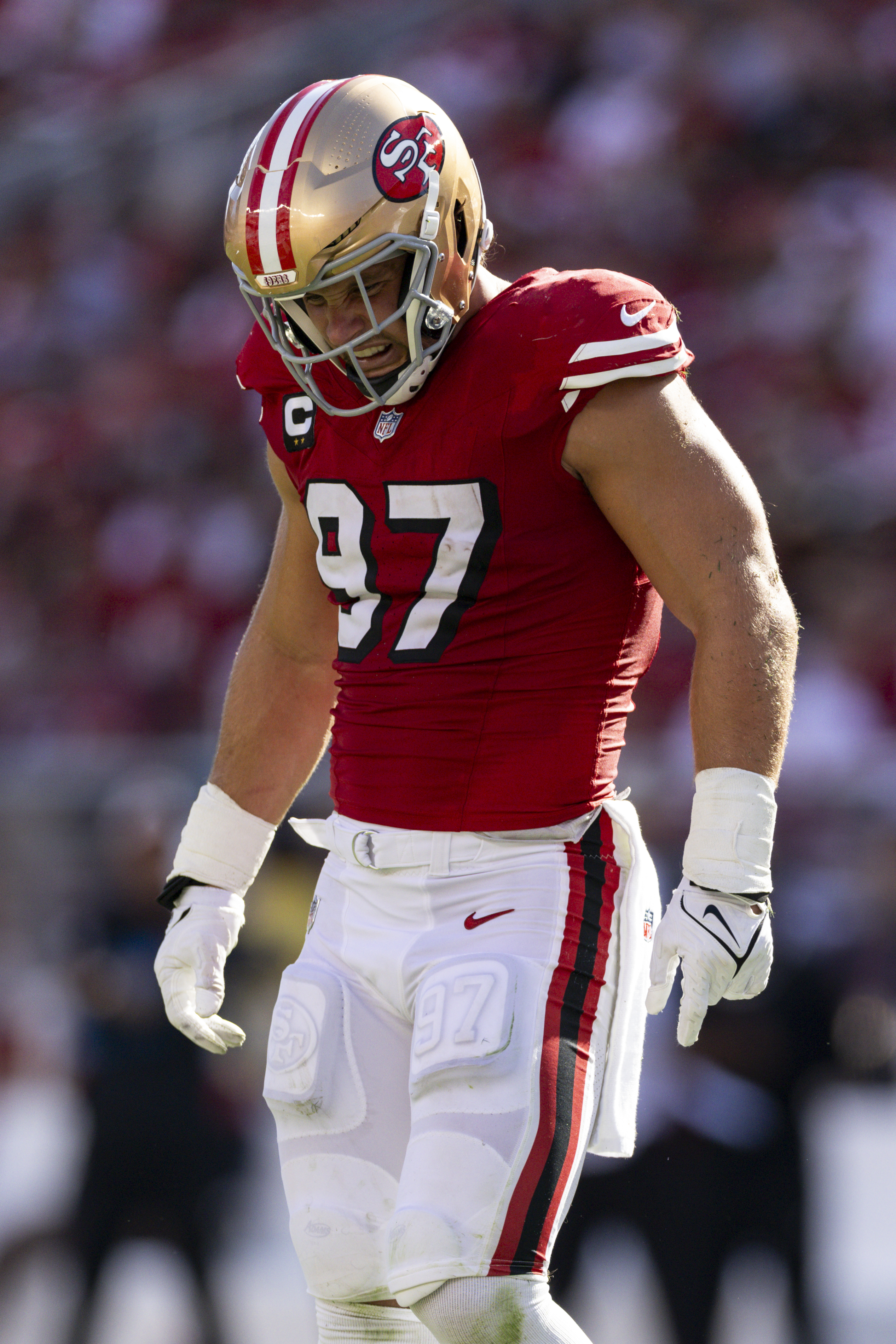 A football player wearing a red jersey and white pants, marked with number 97, stands on the field in action, sporting a gold helmet with a team logo.