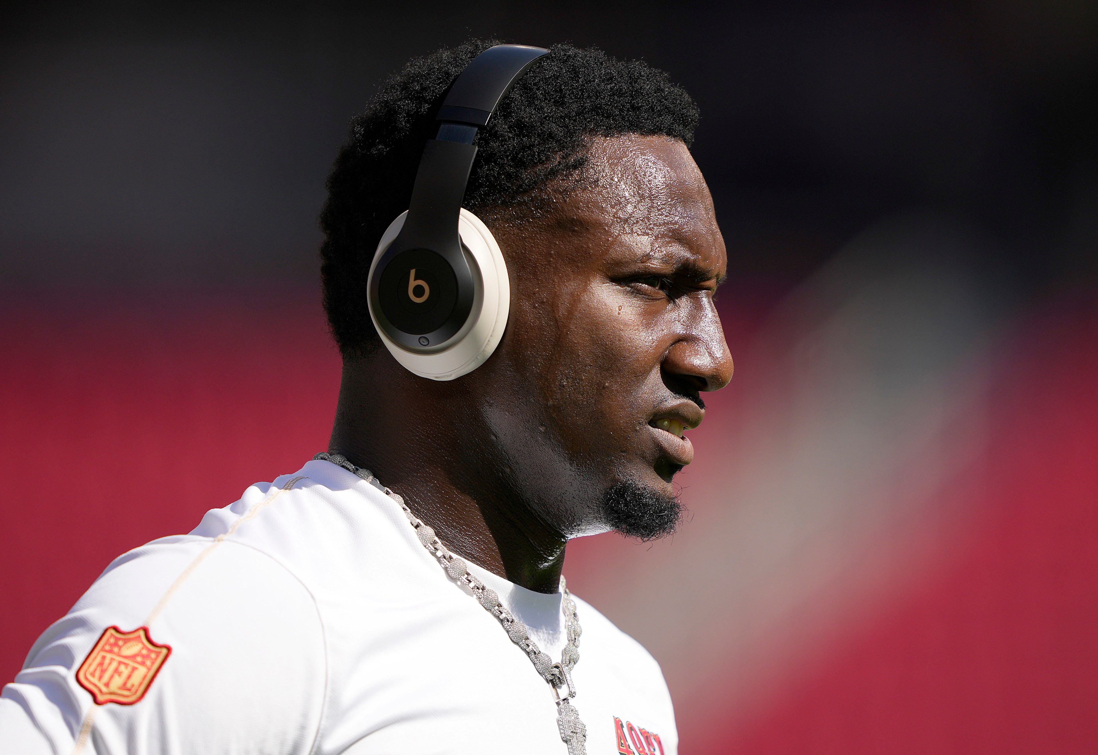 A man wearing large black headphones is outdoors. He's in profile view, wearing a white shirt with an NFL patch, focused and seemingly in mid-motion.