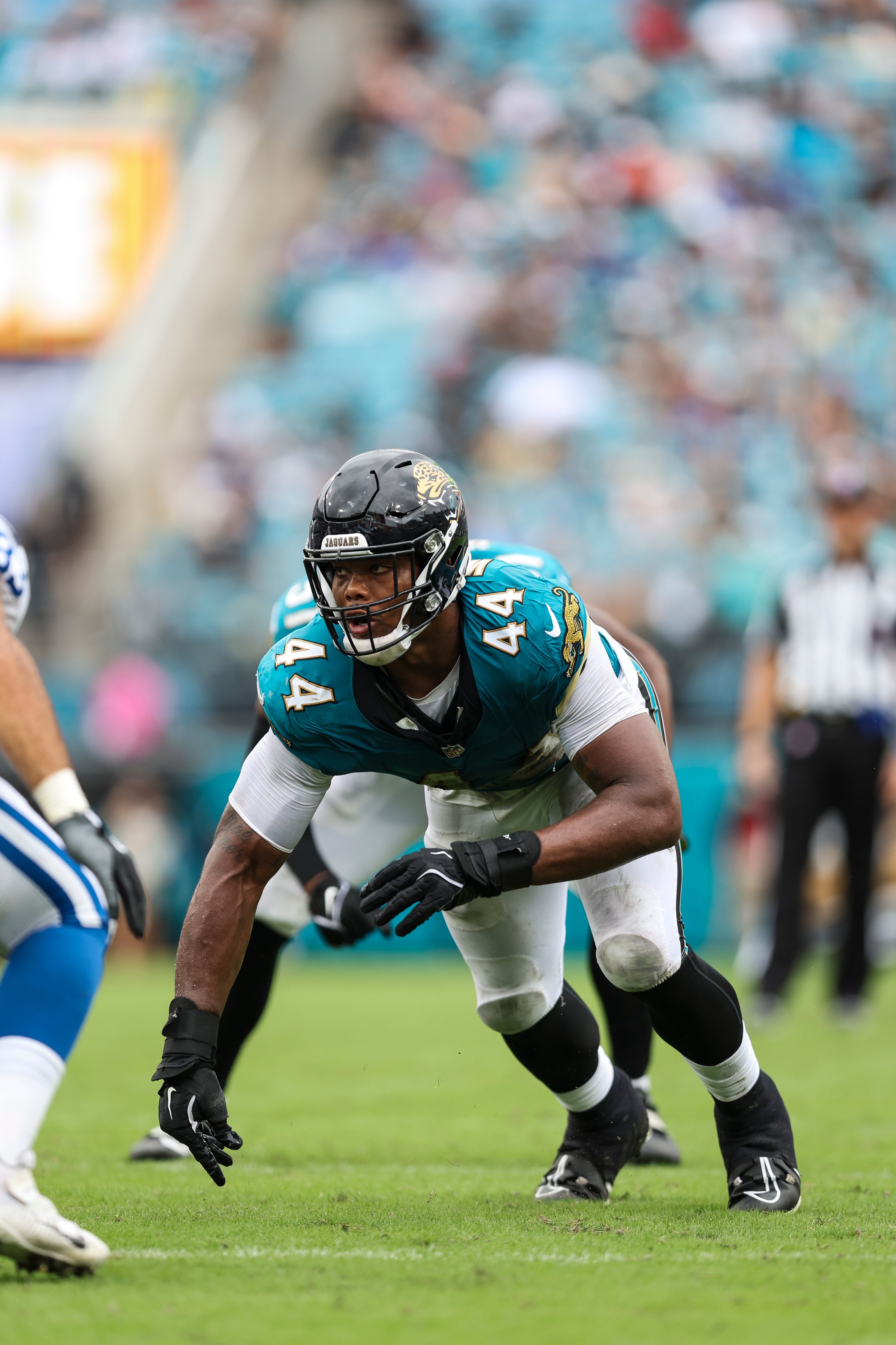 A football player wearing a teal and white uniform with the number 44 and a black helmet crouches on a football field, ready to engage in play.