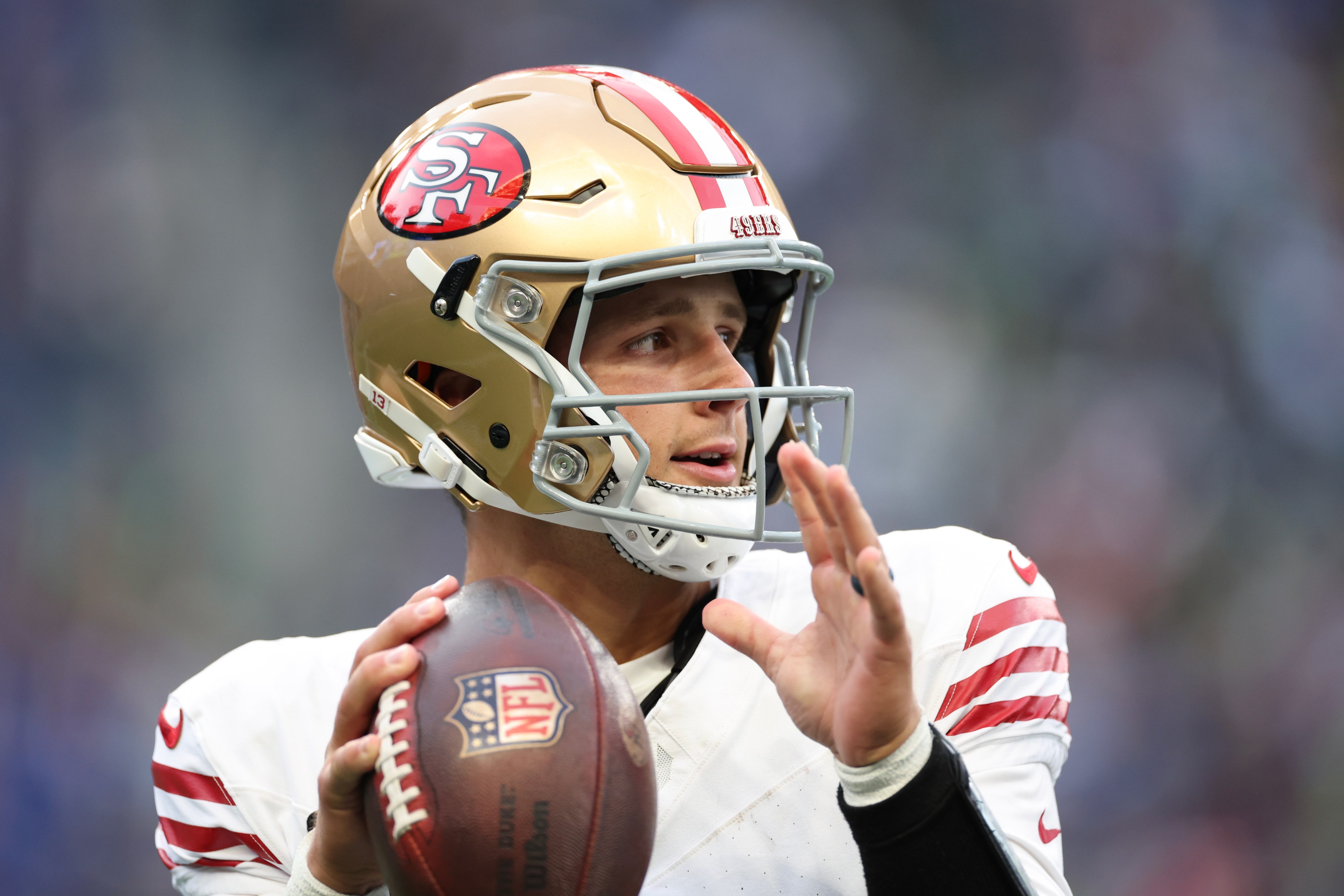 A football player in a gold helmet and white jersey is looking to throw a ball. He wears a helmet with a red and gold logo and has his arm cocked to pass.