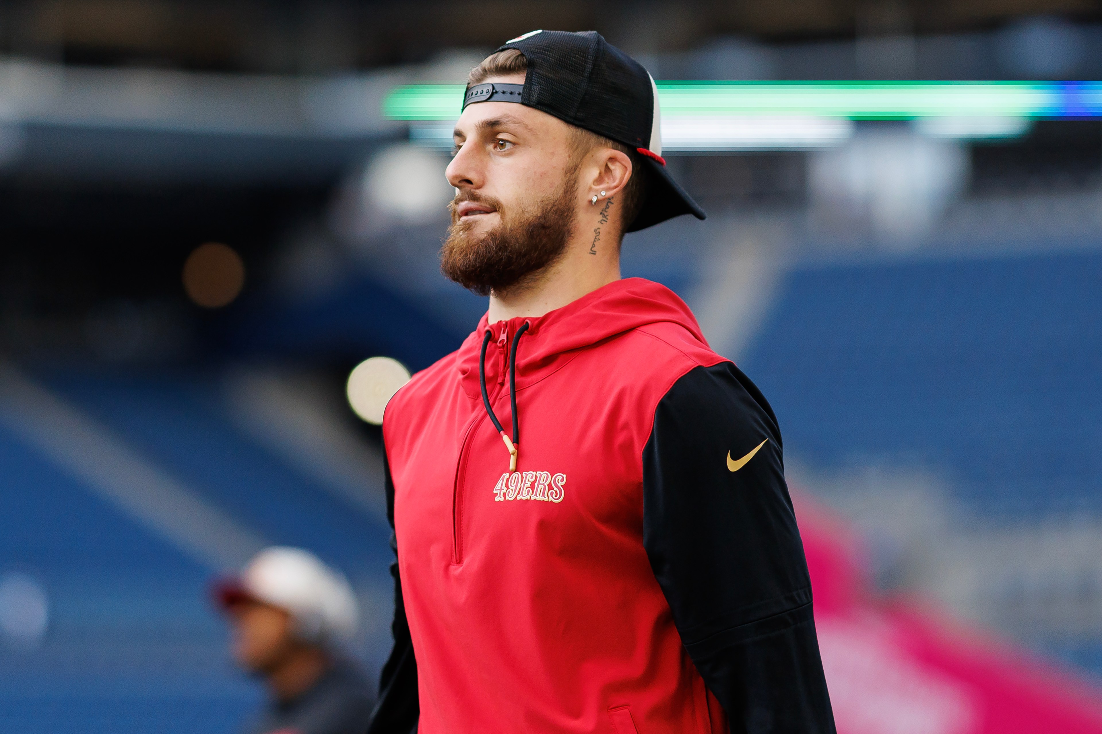 A man with a beard is wearing a backward black cap and a red "49ers" hoodie. He stands in a stadium with unfocused seats and lights in the background.