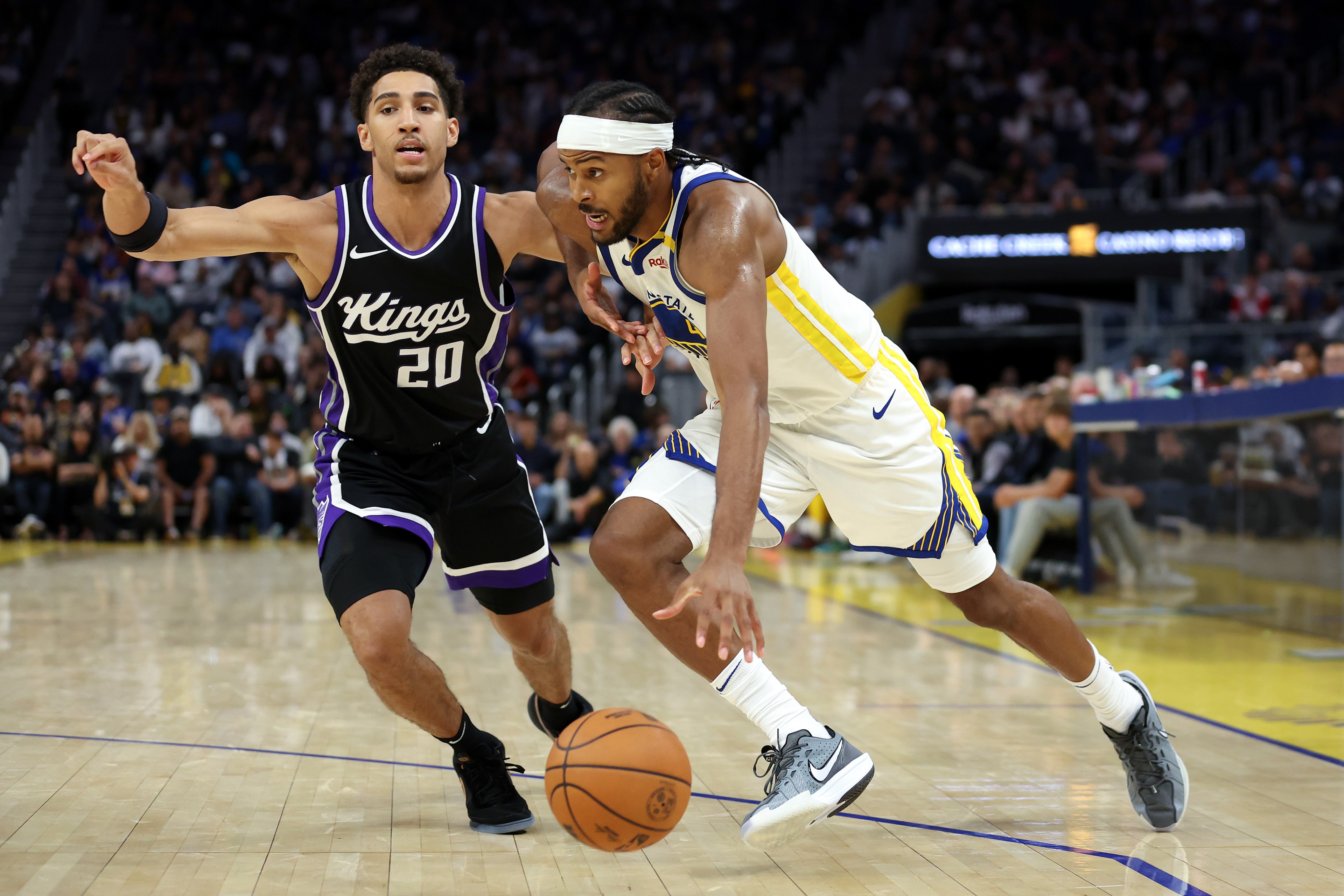 Two basketball players are on a court. One in a black Kings jersey defends as the other in a white Warriors jersey dribbles the ball aggressively.