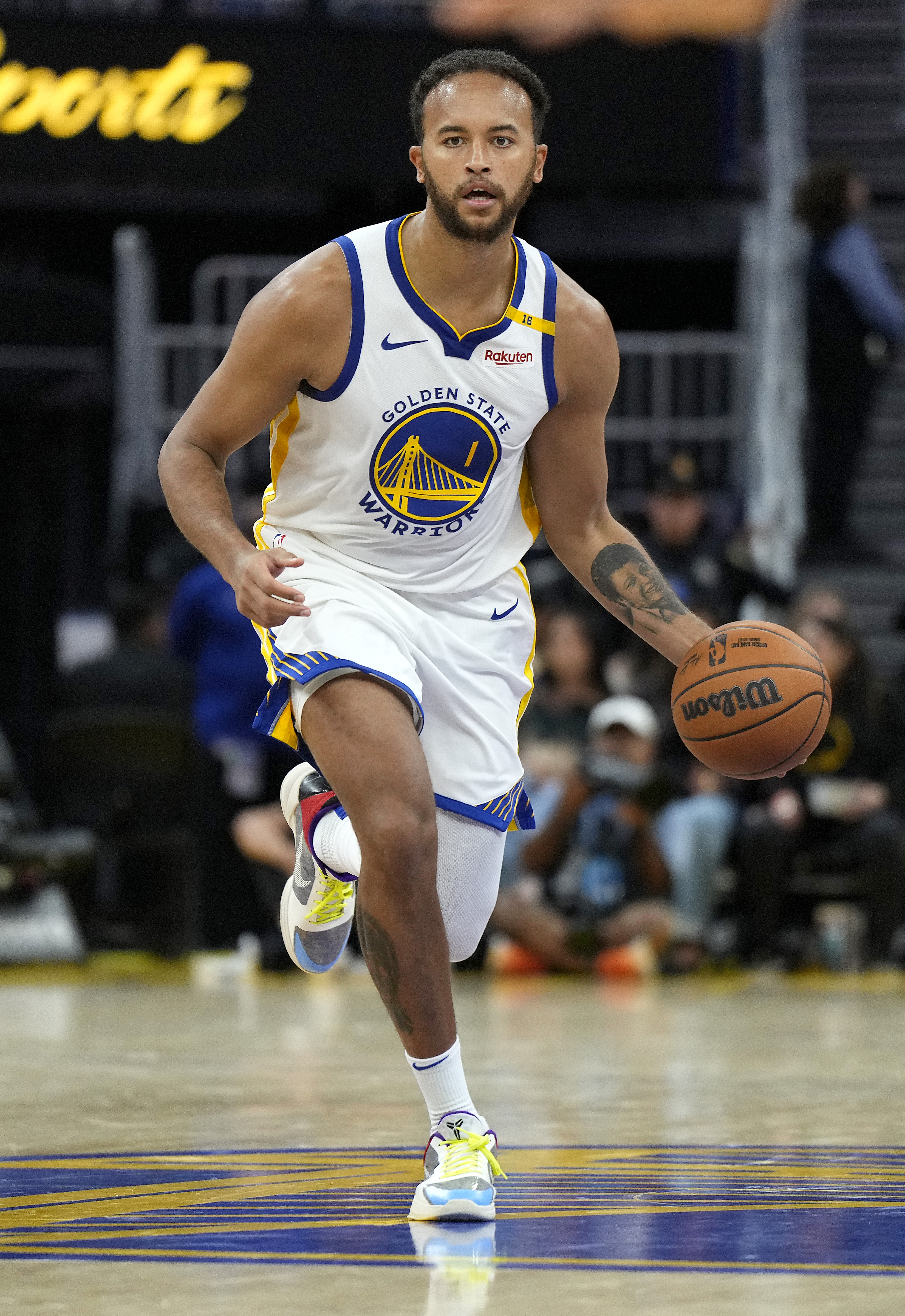 A basketball player in a Golden State Warriors uniform dribbles a basketball on the court, mid-motion, focused. The court's logo is visible underfoot.