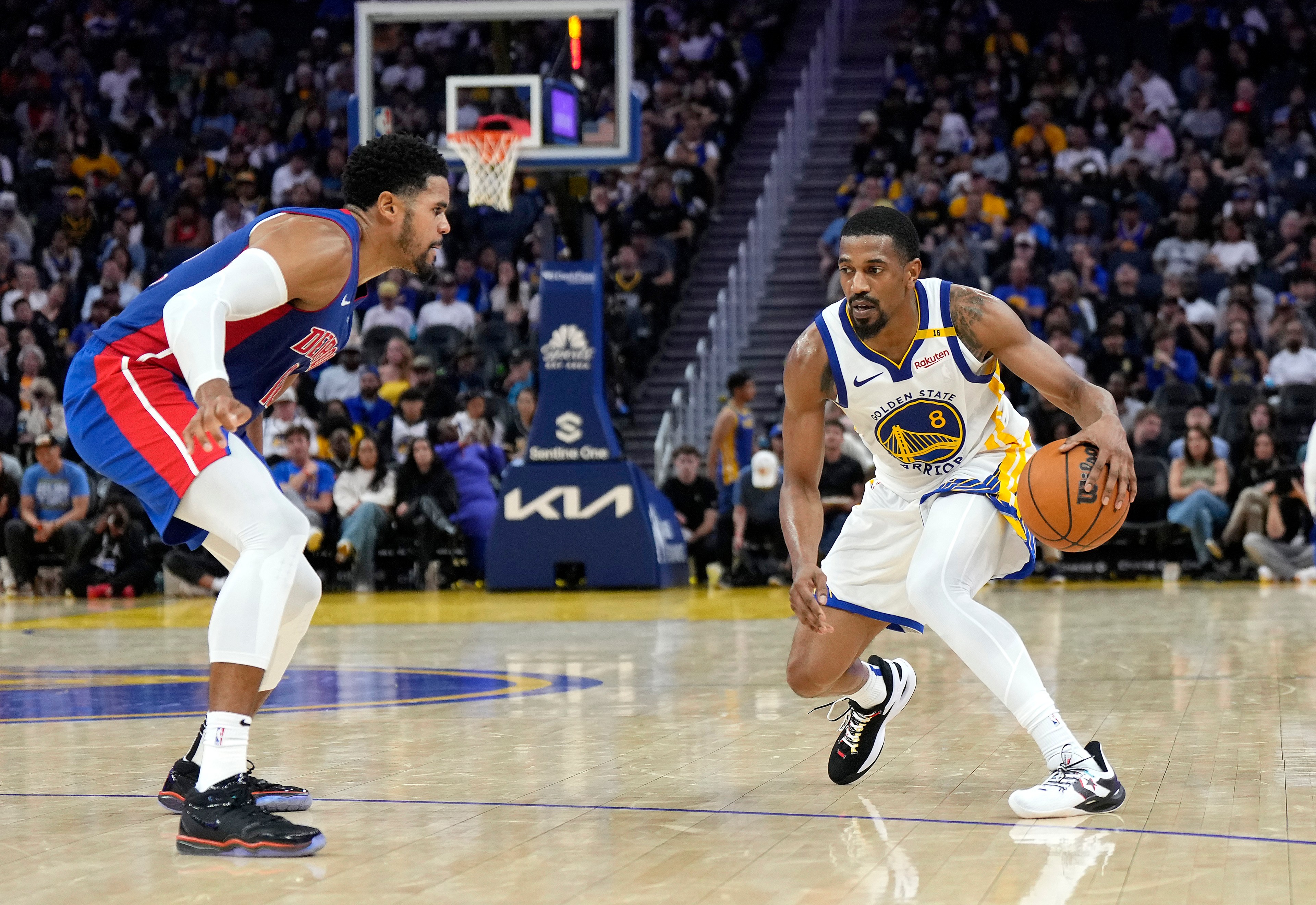 A basketball player in a Golden State Warriors uniform dribbles the ball while being guarded by a player in a red and blue uniform on a well-lit court.