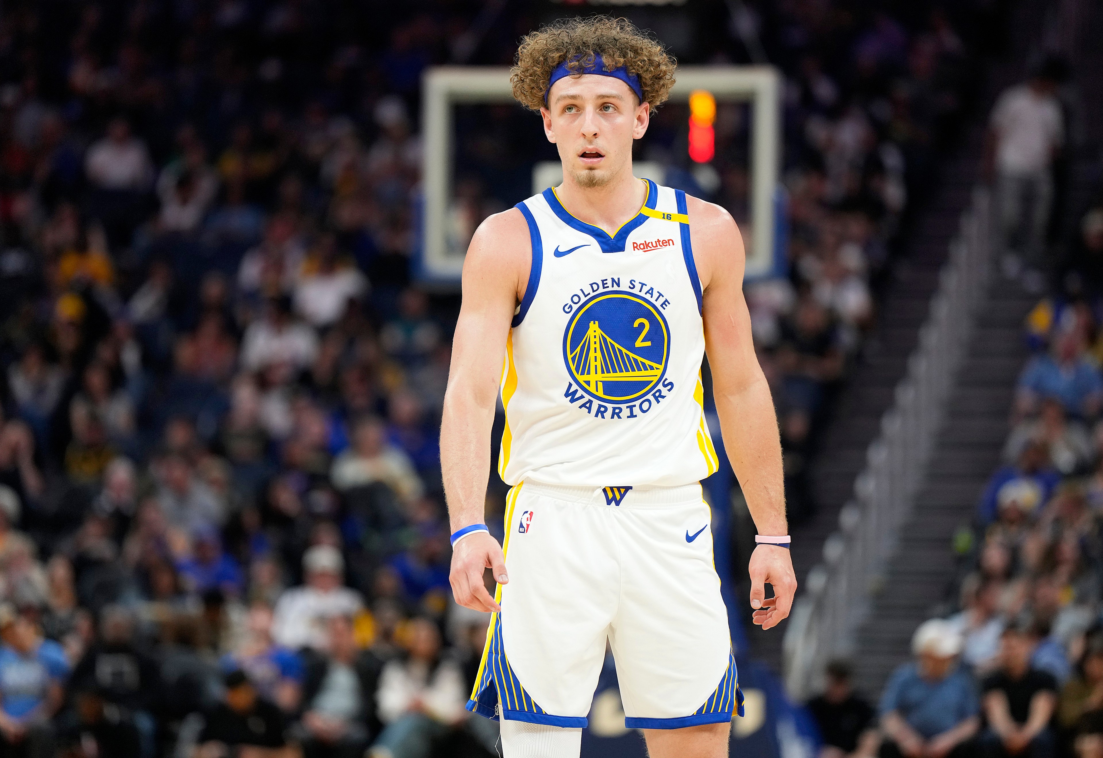 A basketball player wearing a Golden State Warriors jersey stands on the court. He has curly hair with a headband, and the background shows blurred spectators.