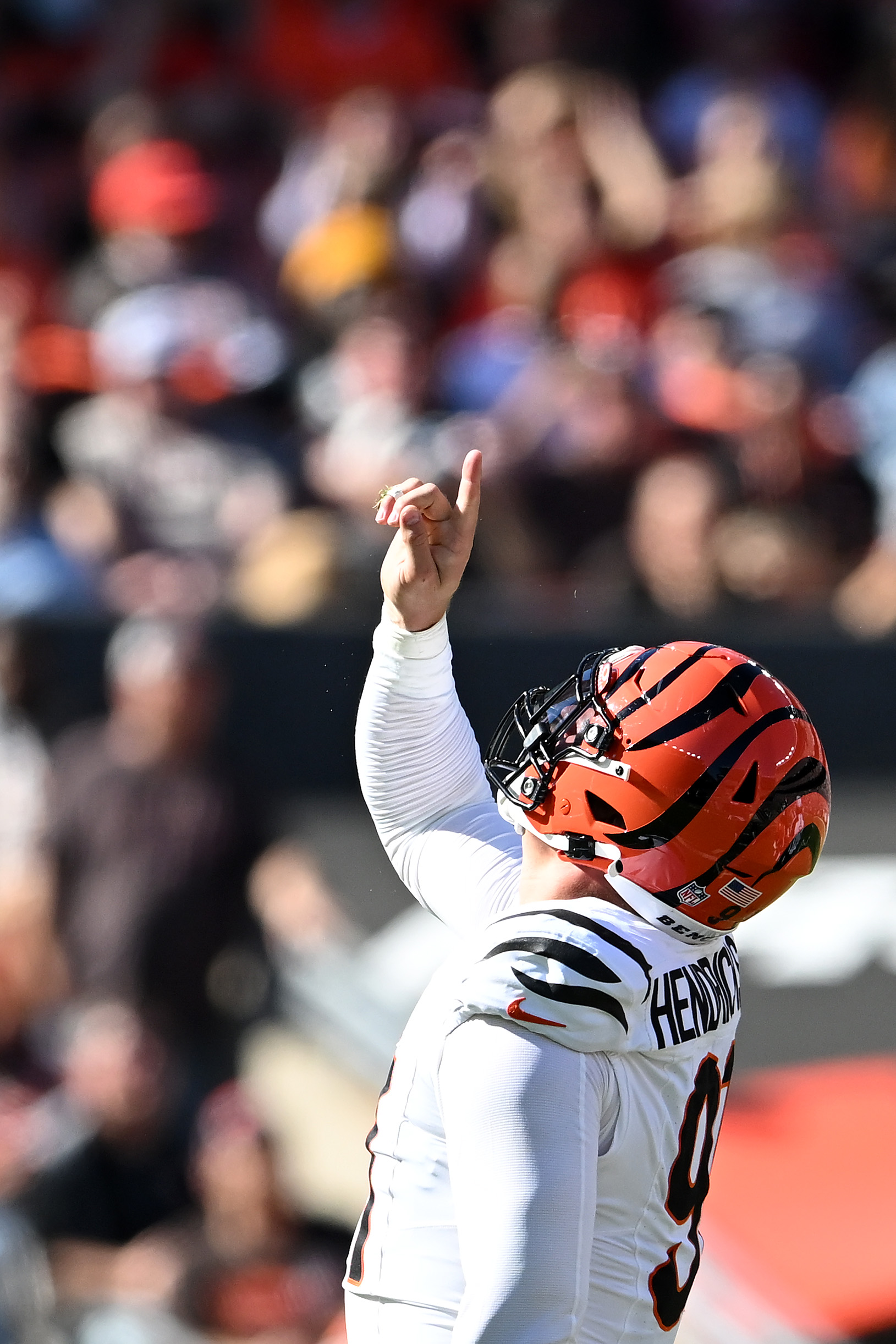 A football player in a white uniform with orange and black accents points upwards. He's wearing a matching helmet, and the crowd is blurred in the background.