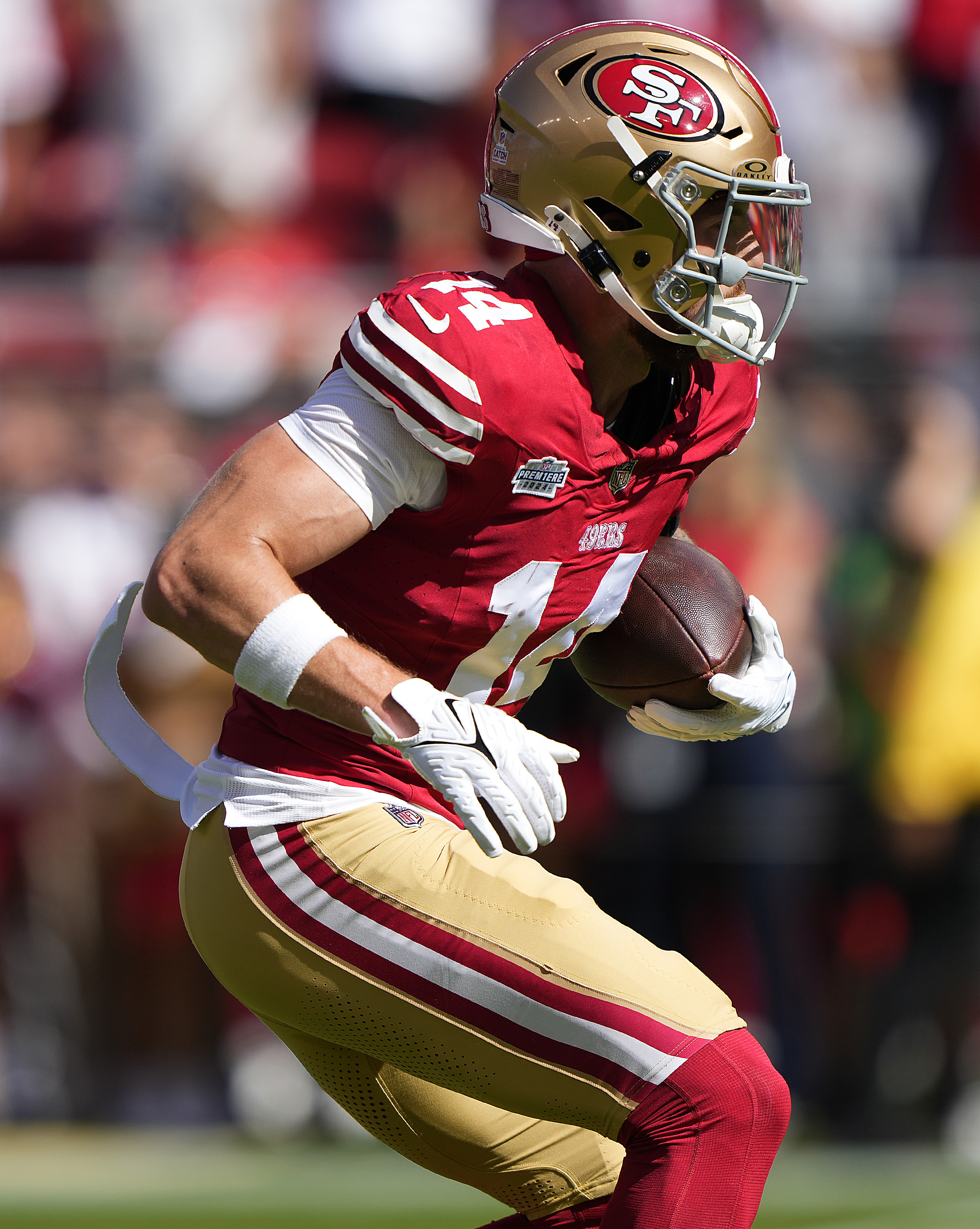 A football player in a red and gold uniform runs with the ball. He wears a helmet with "SF" on it and white gloves, focused and in action on the field.