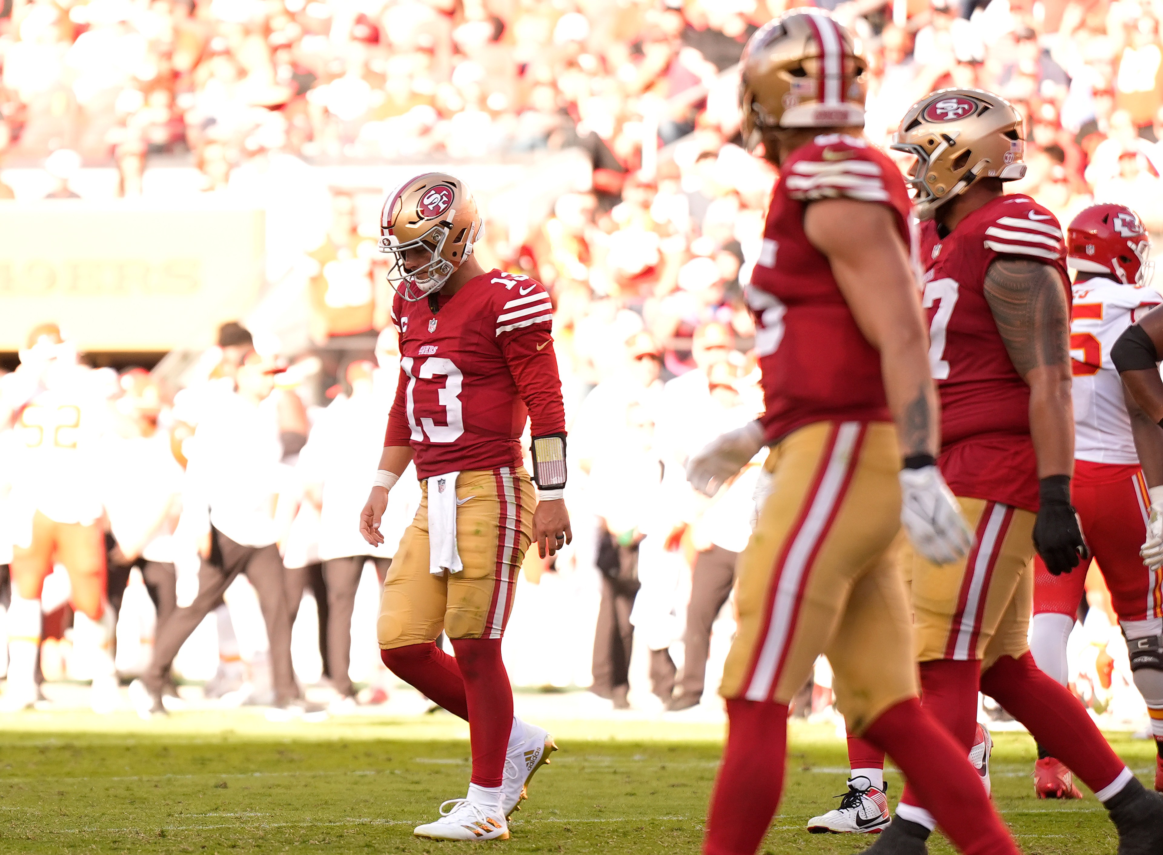 The image shows football players in red and gold uniforms on a field, with one player looking down. The background is filled with a blurred crowd.