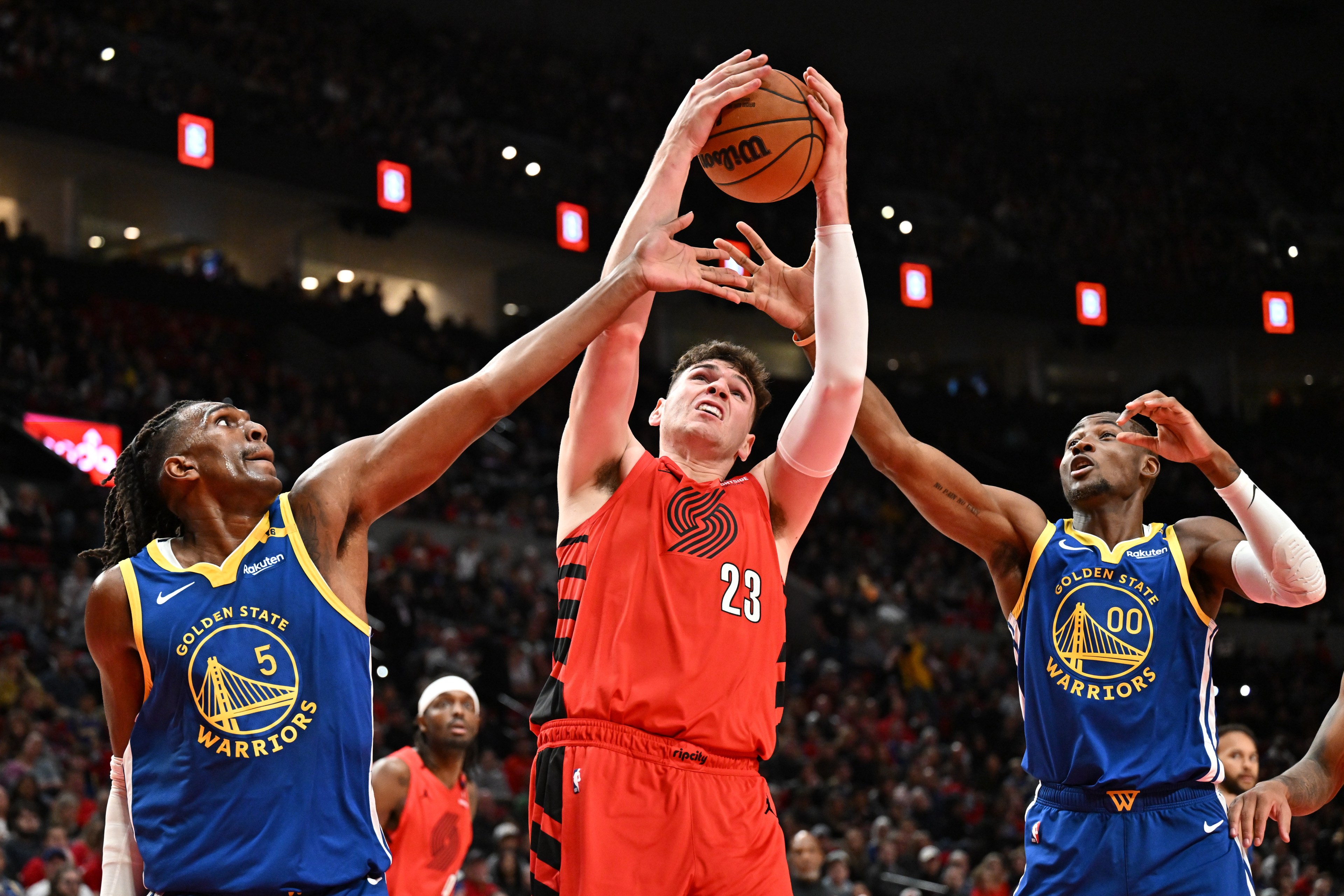 Three basketball players are reaching for the ball mid-air. Two are in blue Golden State Warriors jerseys, while the player in the middle wears a red jersey.