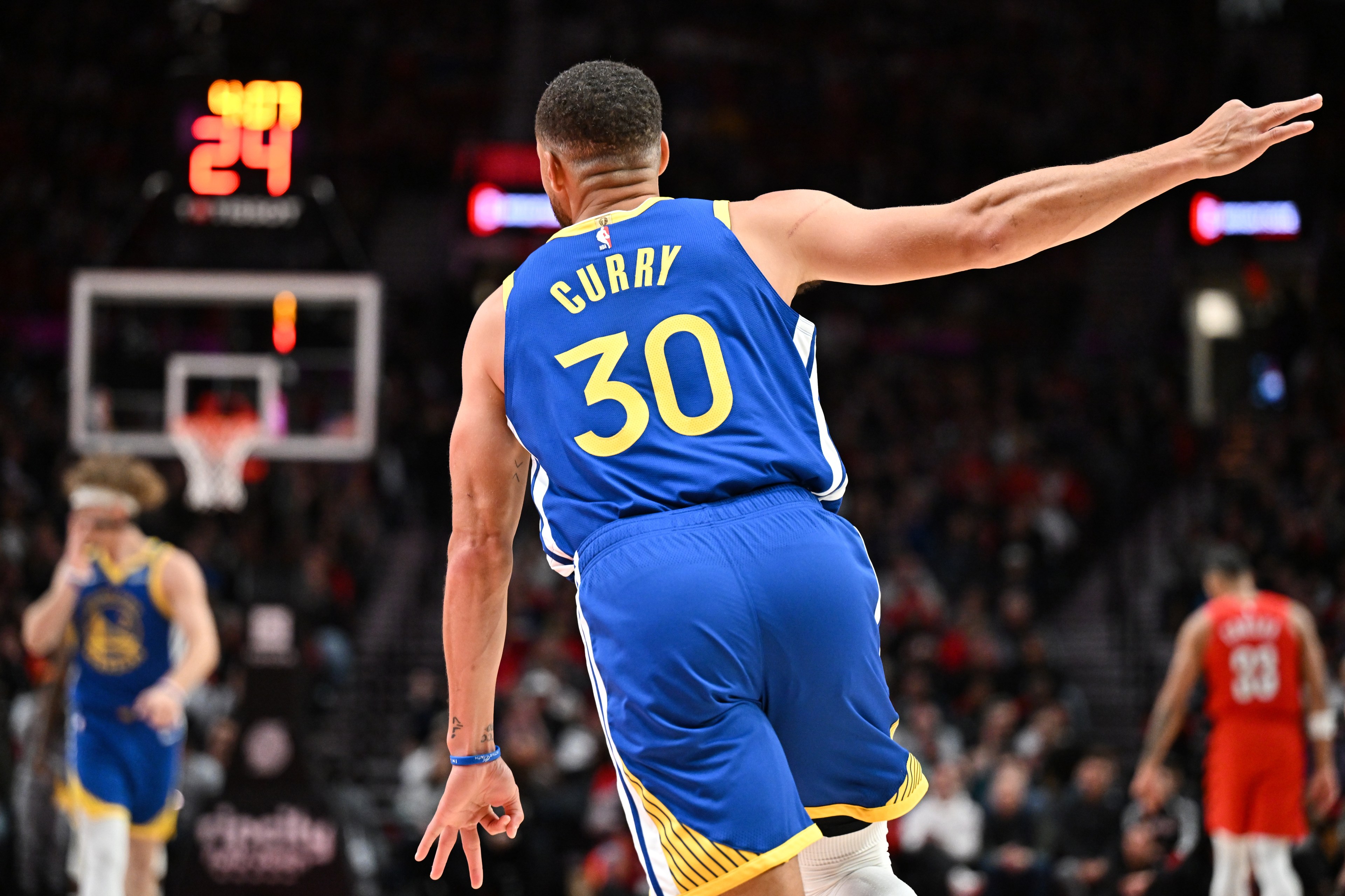 A basketball player wearing a blue jersey with number 30 raises one arm, facing the hoop. The background shows another player and a digital shot clock.