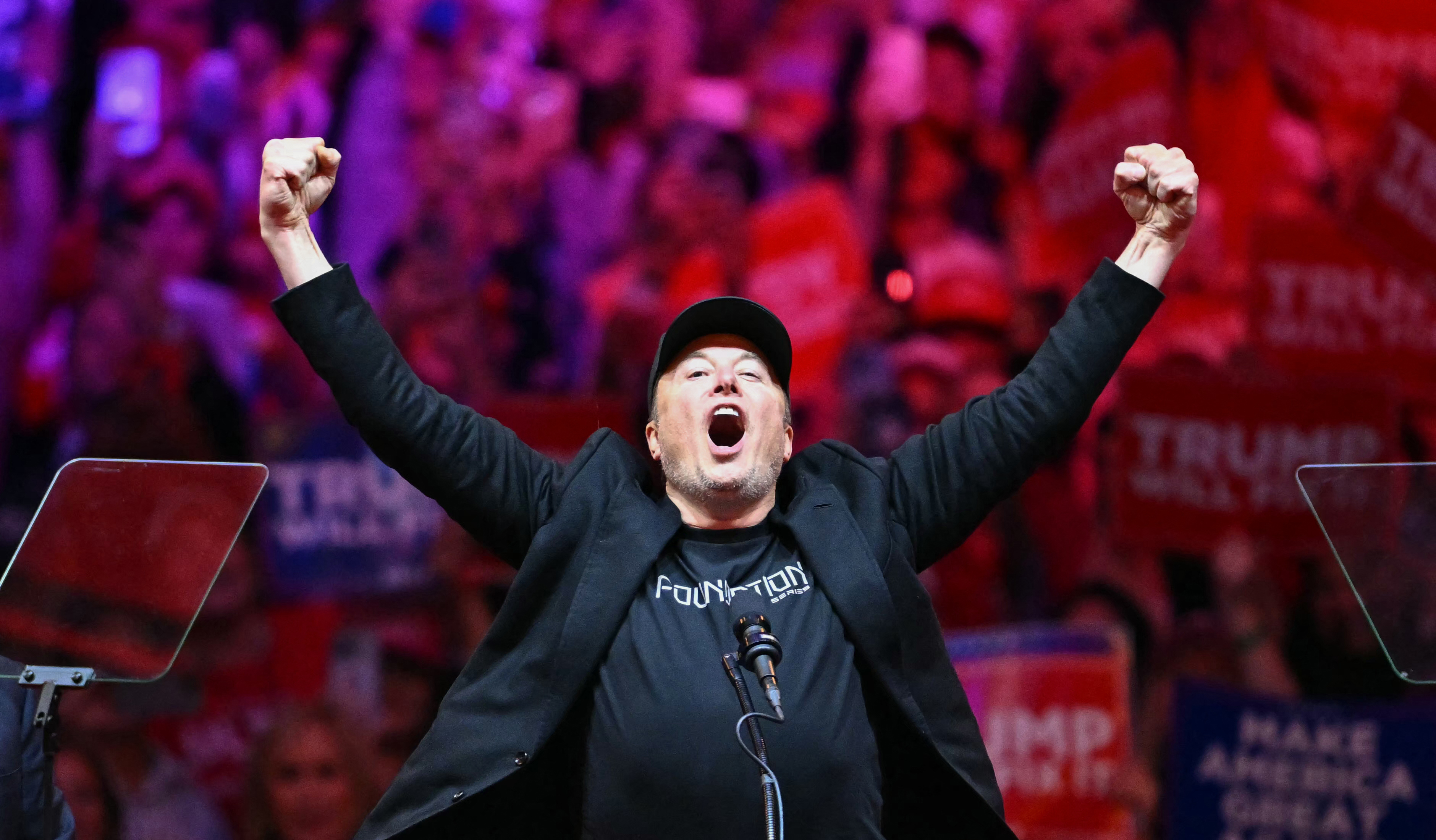 A person in a black outfit and cap stands with arms raised, shouting into a microphone. Behind them, a crowd holds red signs in a vibrant, colorful setting.