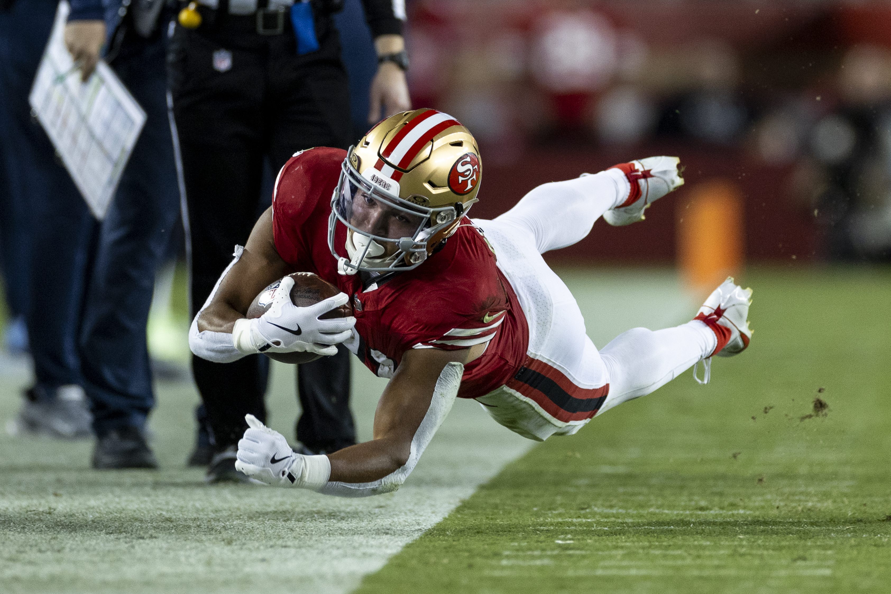 A football player in a red and gold uniform is diving near the sideline, holding the ball tightly, with one foot off the ground and focus in his eyes.