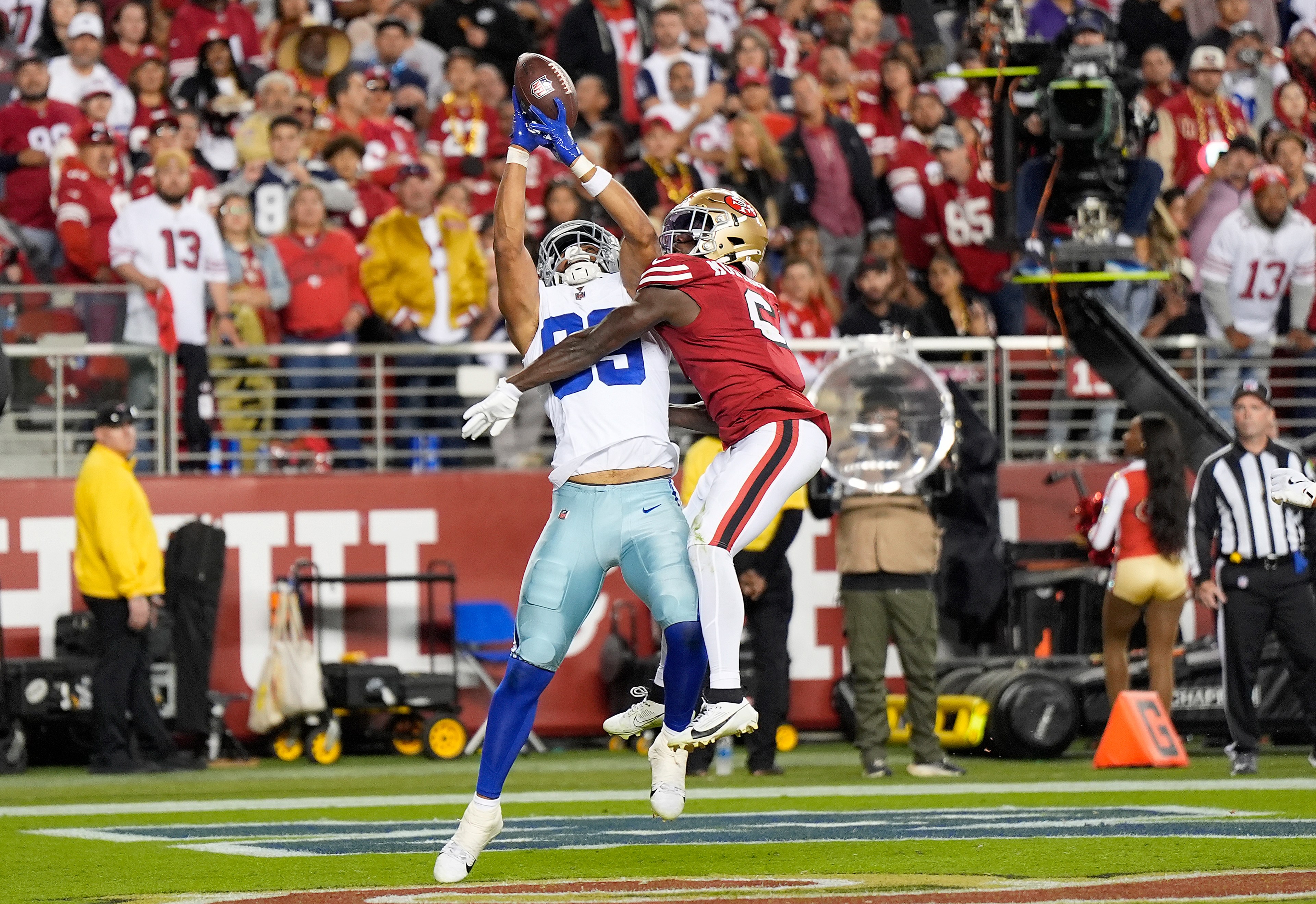 Two football players leap for the ball during a game. One wears a white and blue uniform, the other red and gold. A crowd watches in the background.