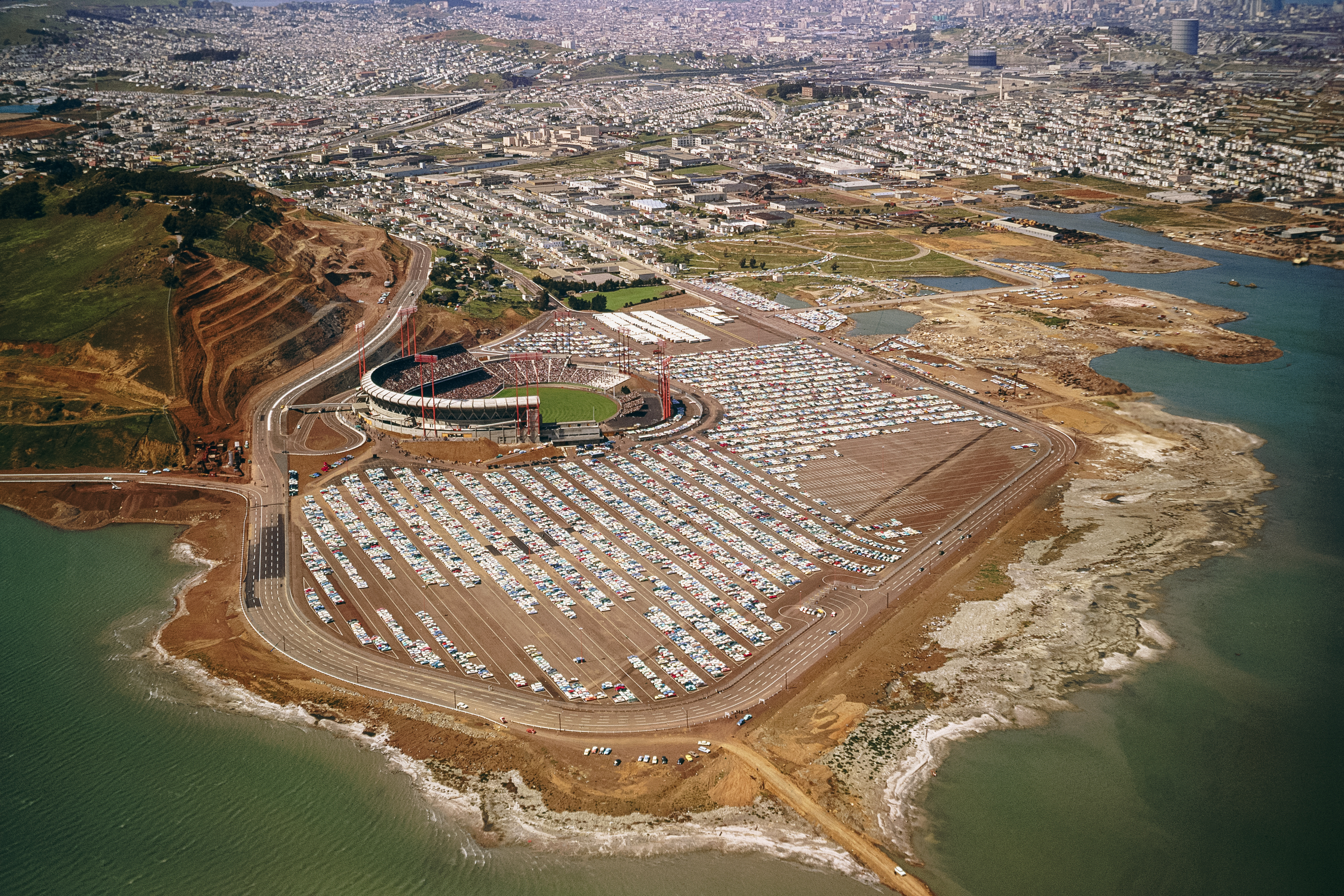 The image shows an aerial view of a stadium with a massive parking lot filled with cars. Surrounding the area are bodies of water and urban developments.