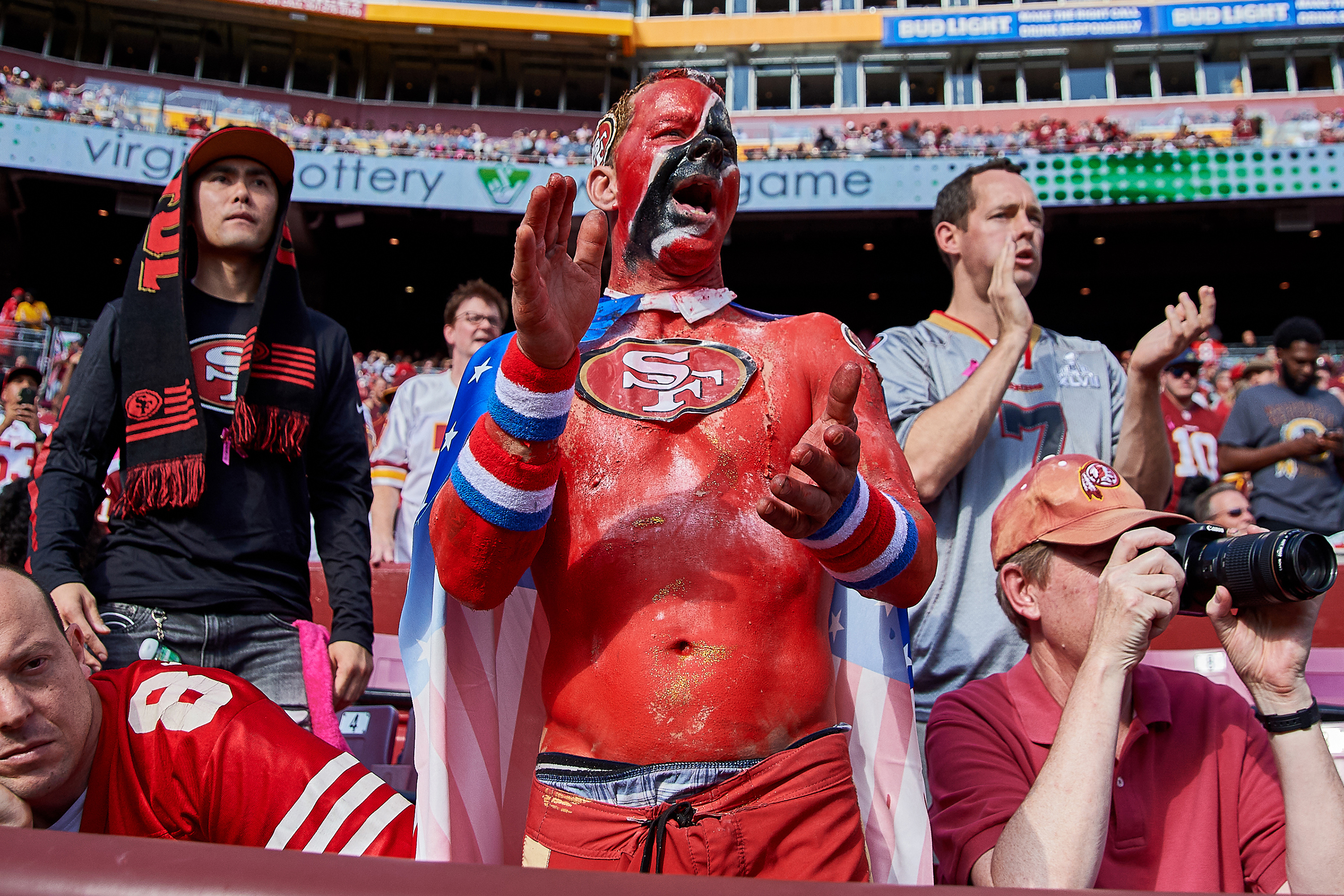 A fan painted in red with a large &quot;SF&quot; logo on his chest is cheering in a stadium, surrounded by other fans, including one taking photos.