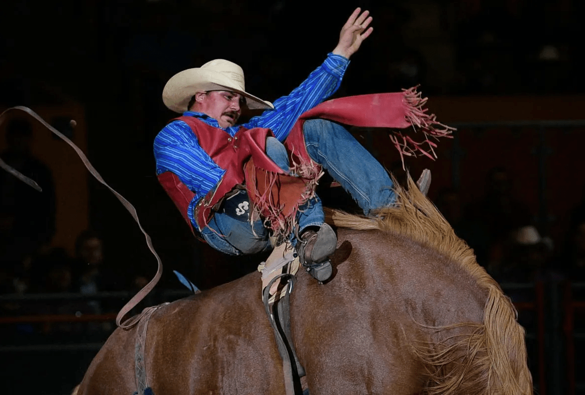 A rodeo cowboy in a blue shirt and red chaps is riding a bucking horse, holding on with one hand raised. He's wearing a cowboy hat and focused on staying on.