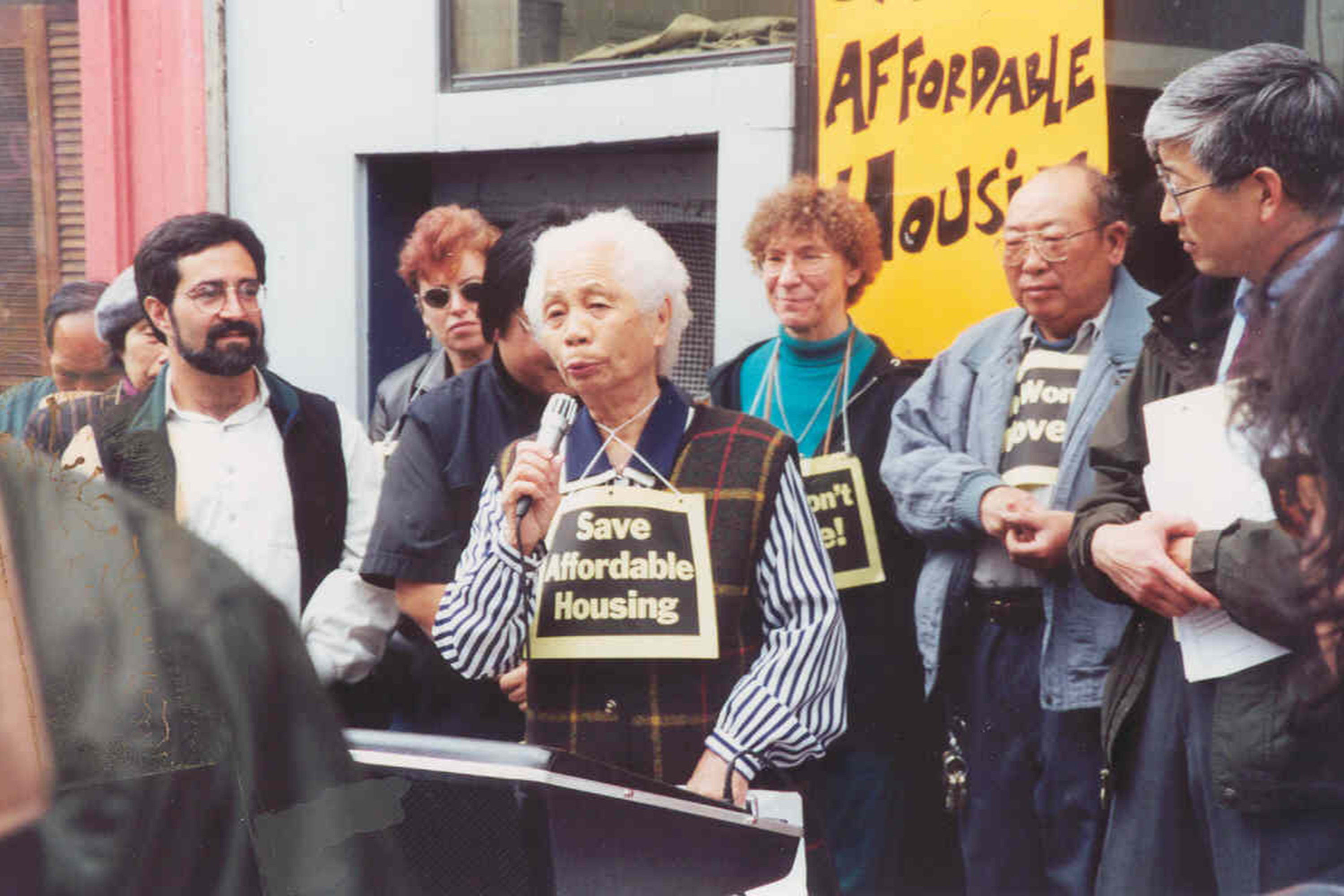 A 1990's photograph of an elderly woman speaking from a microphone outside a home with protesters and Aaron Peskin standing around her.