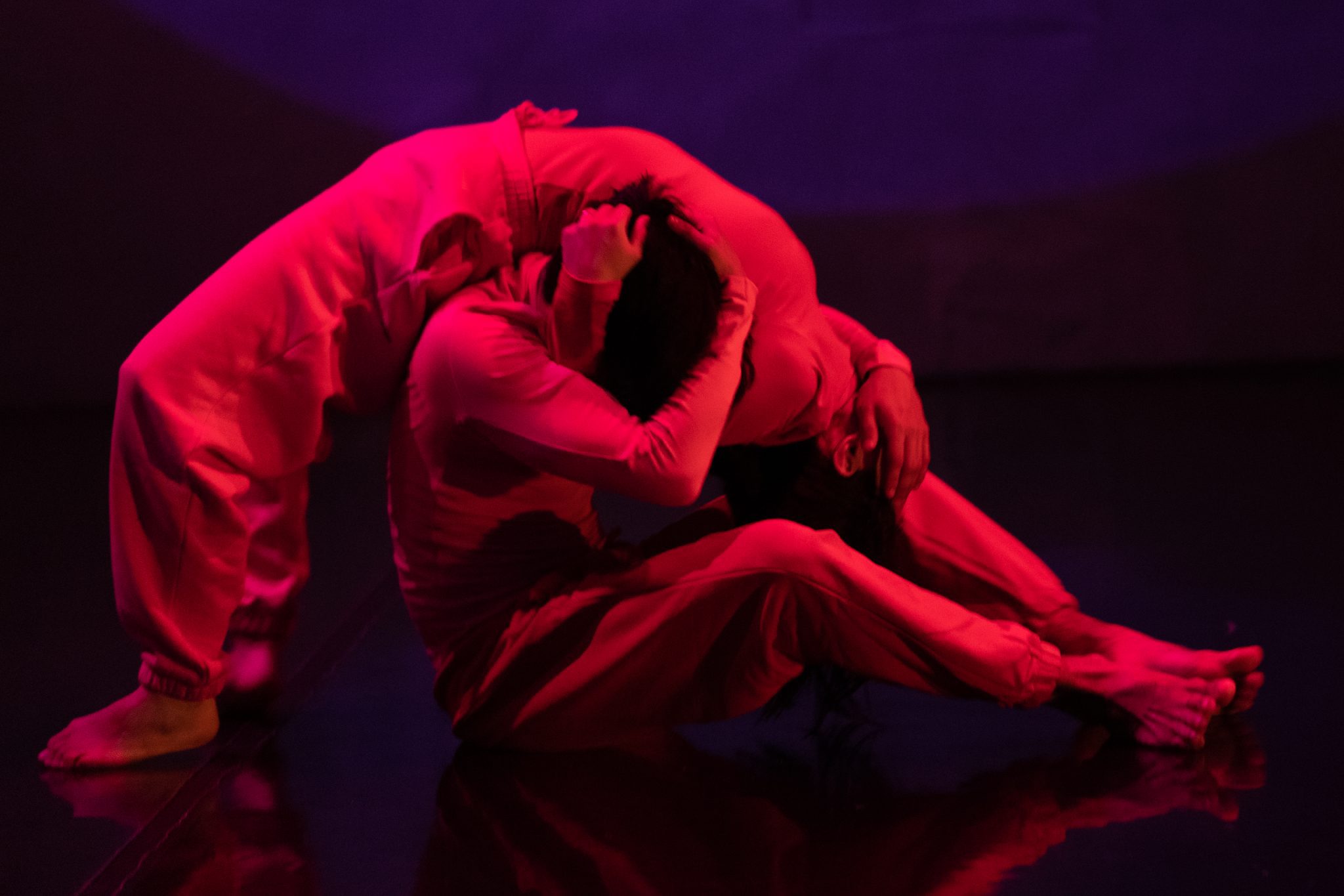 Two dancers in red costumes intertwine on a dimly lit stage with dramatic red lighting, creating a fluid, expressive pose full of emotion and intensity.