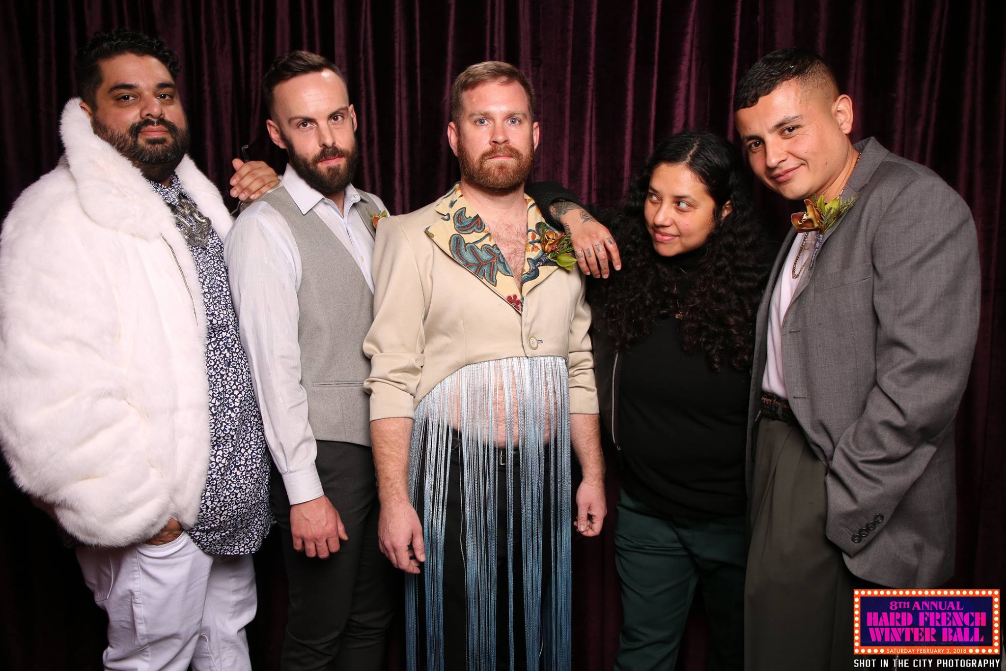 Five people pose together against a dark curtain. They wear a mix of formal and creative outfits, including a light fur coat, a fringed jacket, and floral embellishments.