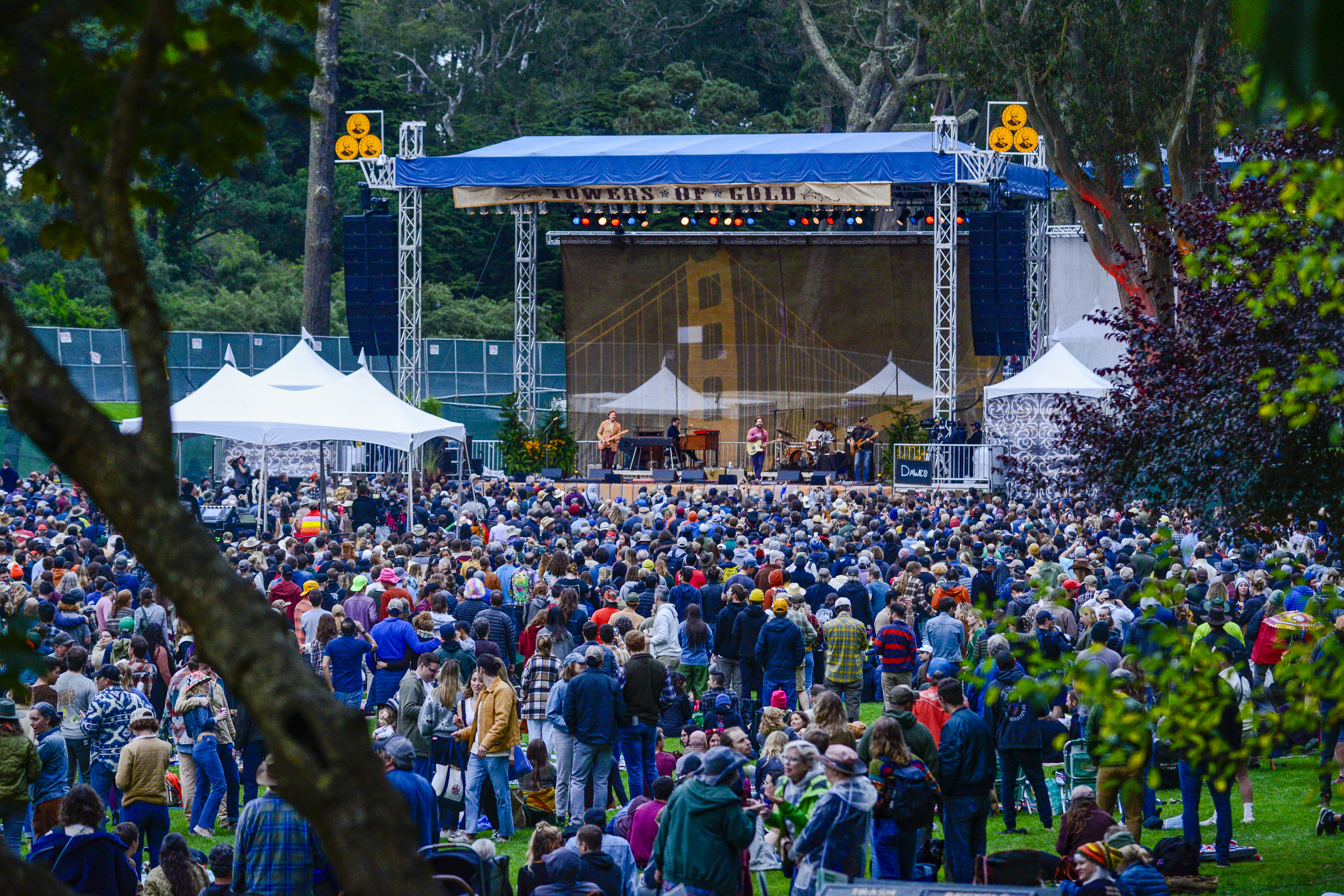 A band performs on a large outdoor stage with a crowd gathered on a grassy area. The backdrop features a bridge design, and trees surround the venue.